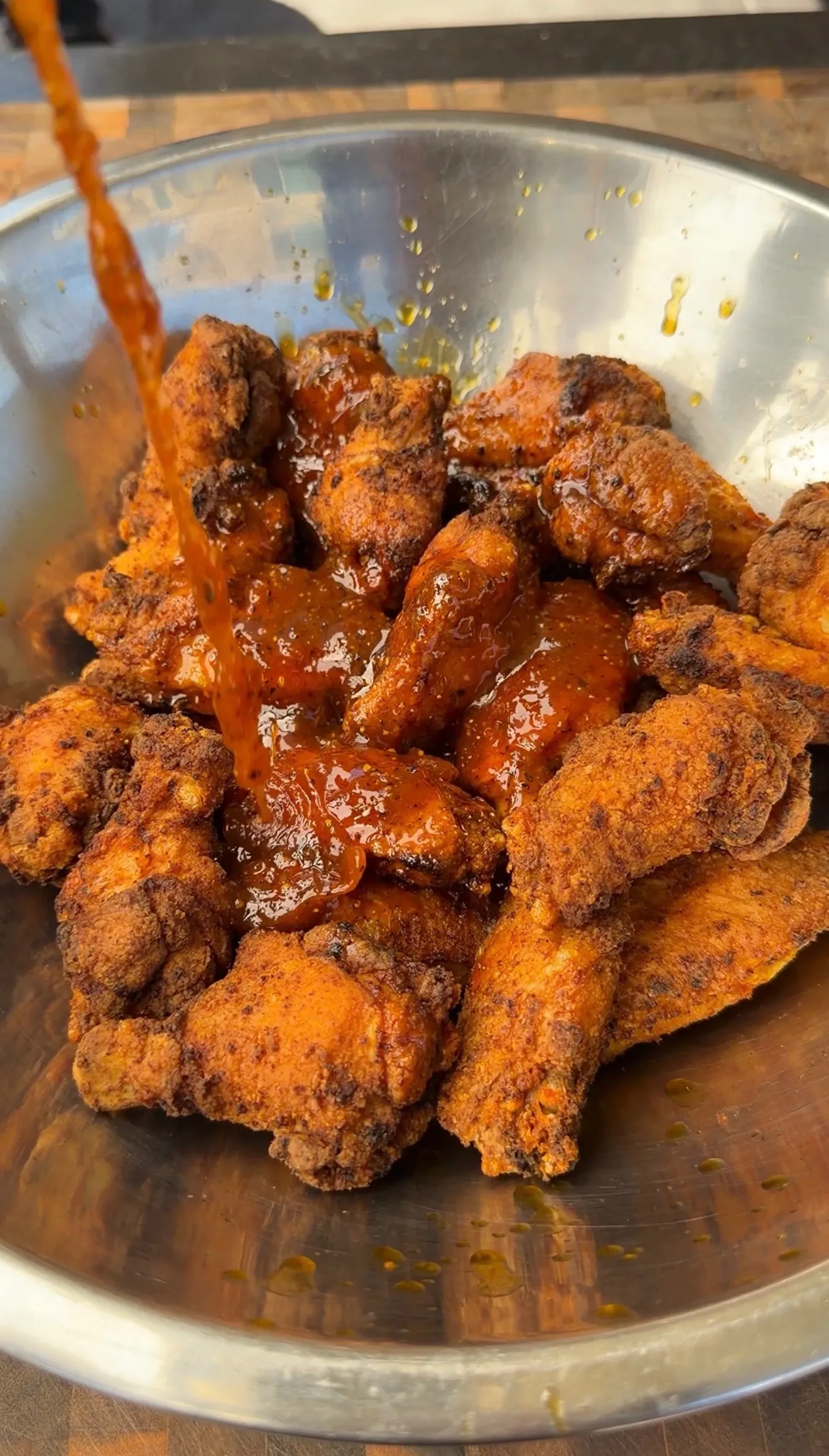 Crispy fried chicken wings in a steel bowl being coated with honey lemon pepper sauce poured from above