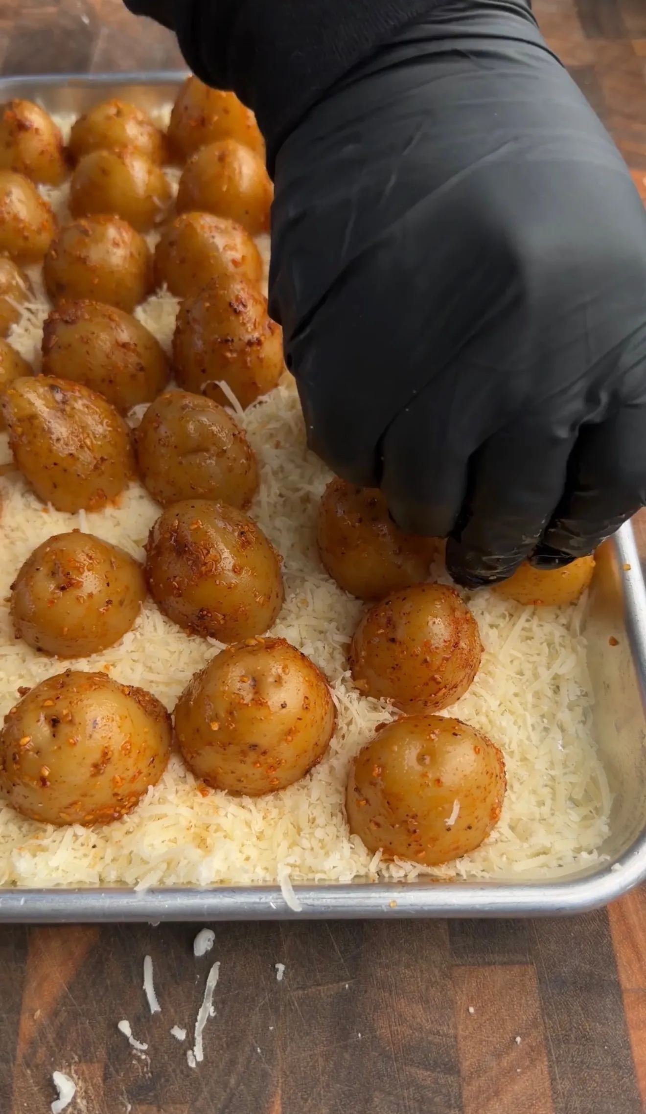 Golden roasted potatoes with melted parmesan cheese on a baking sheet, being prepared with black gloved hand