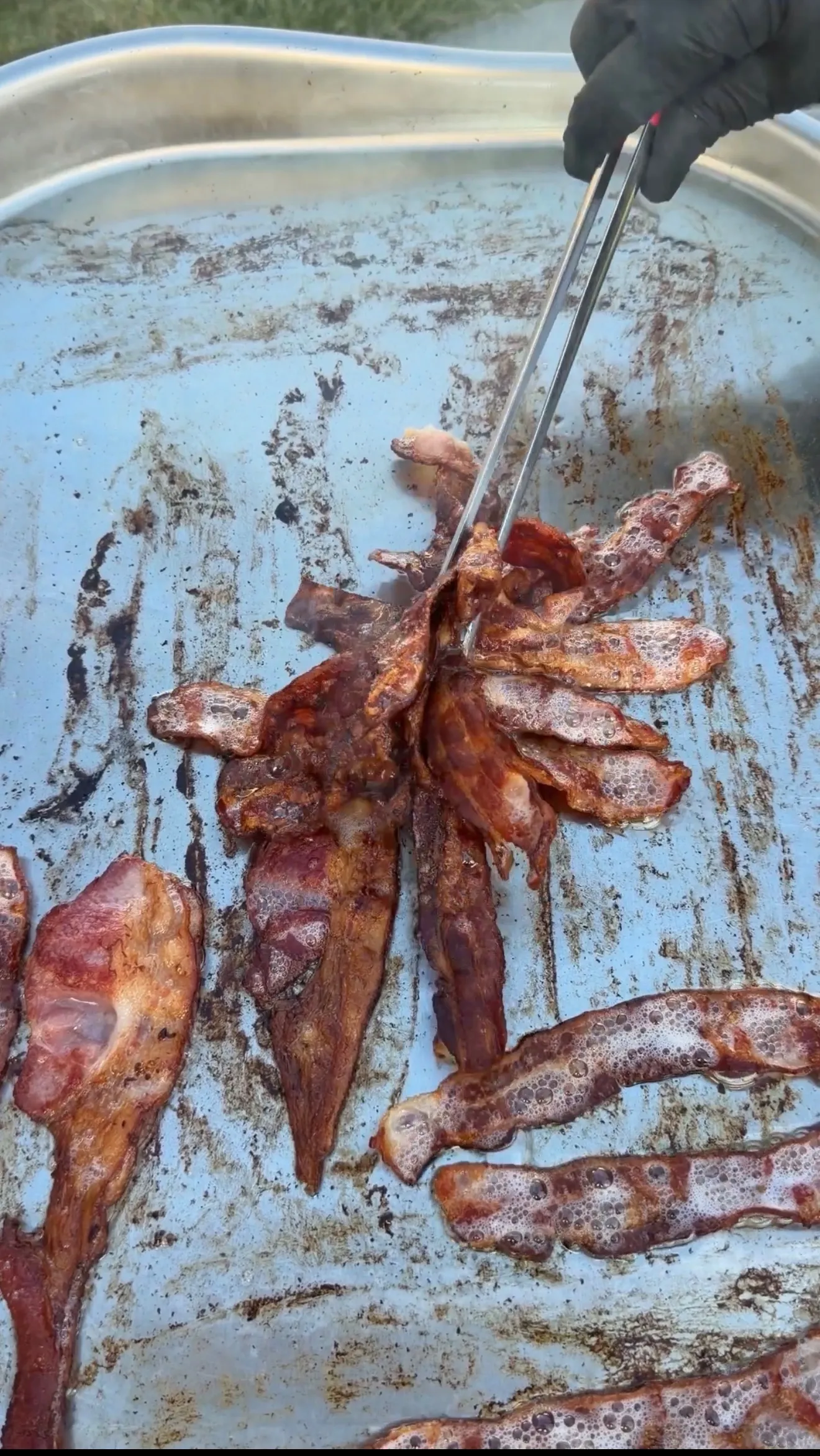 Crispy smoked bacon strips being tossed with tongs on a seasoned flat-top griddle