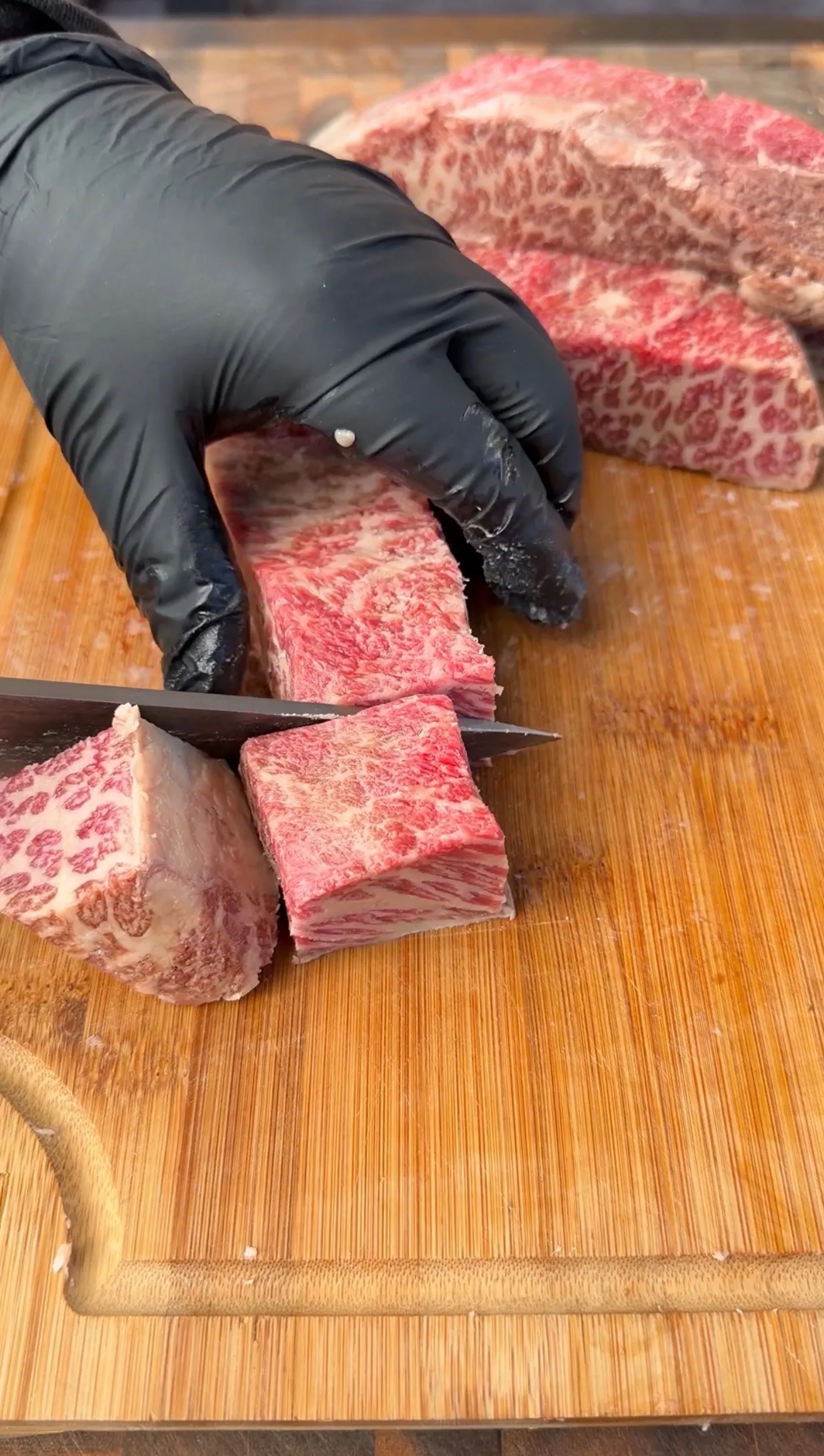 Gloved hands cutting marbled beef short ribs into cubes on a wooden cutting board with sharp knife