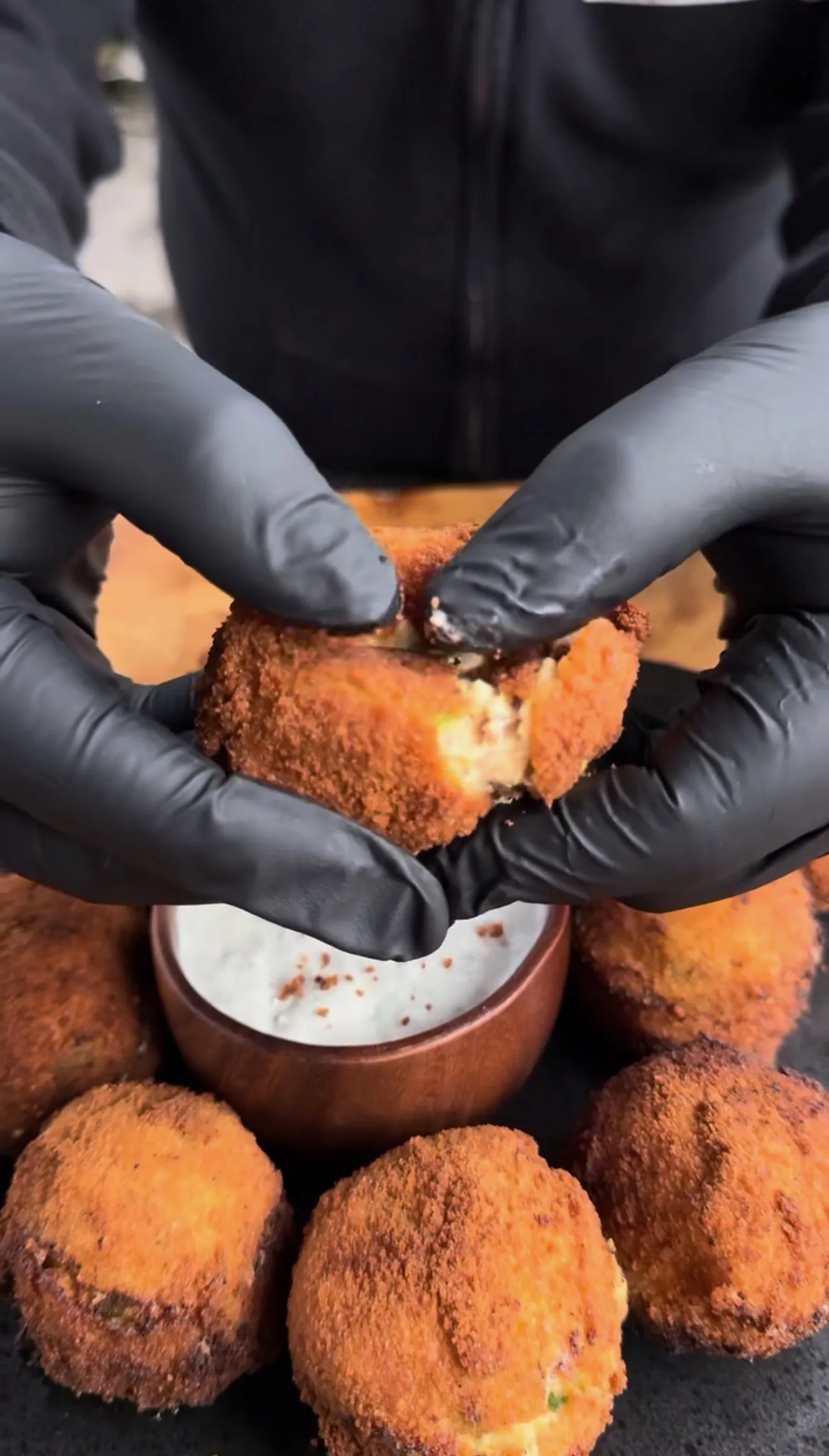 Hands in black gloves breaking open a crispy deep fried jalapeño popper bite over a creamy white dipping sauce
