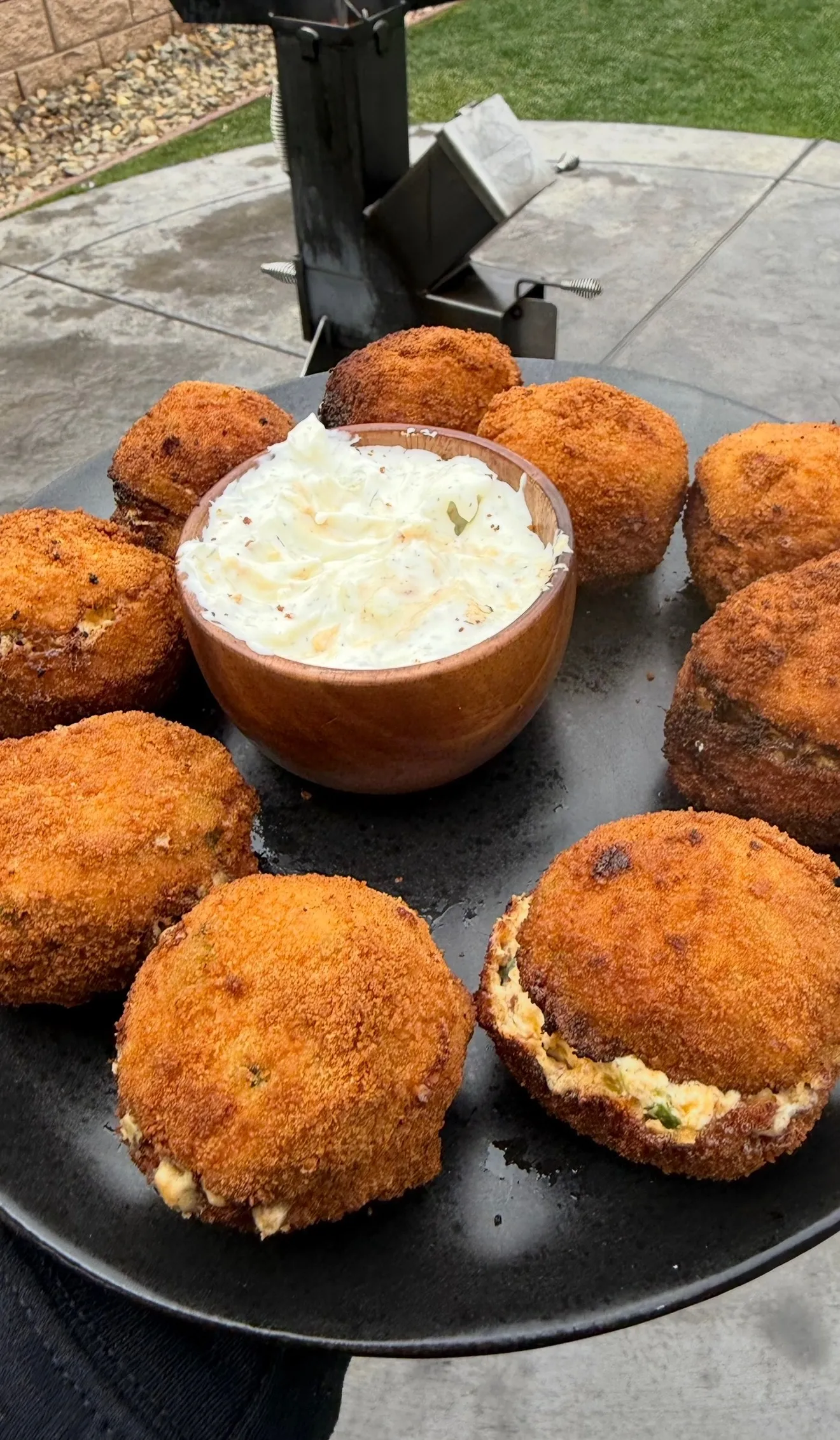 Golden fried jalapeño popper bites arranged on a dark plate around a bowl of creamy dipping sauce, one bitten open