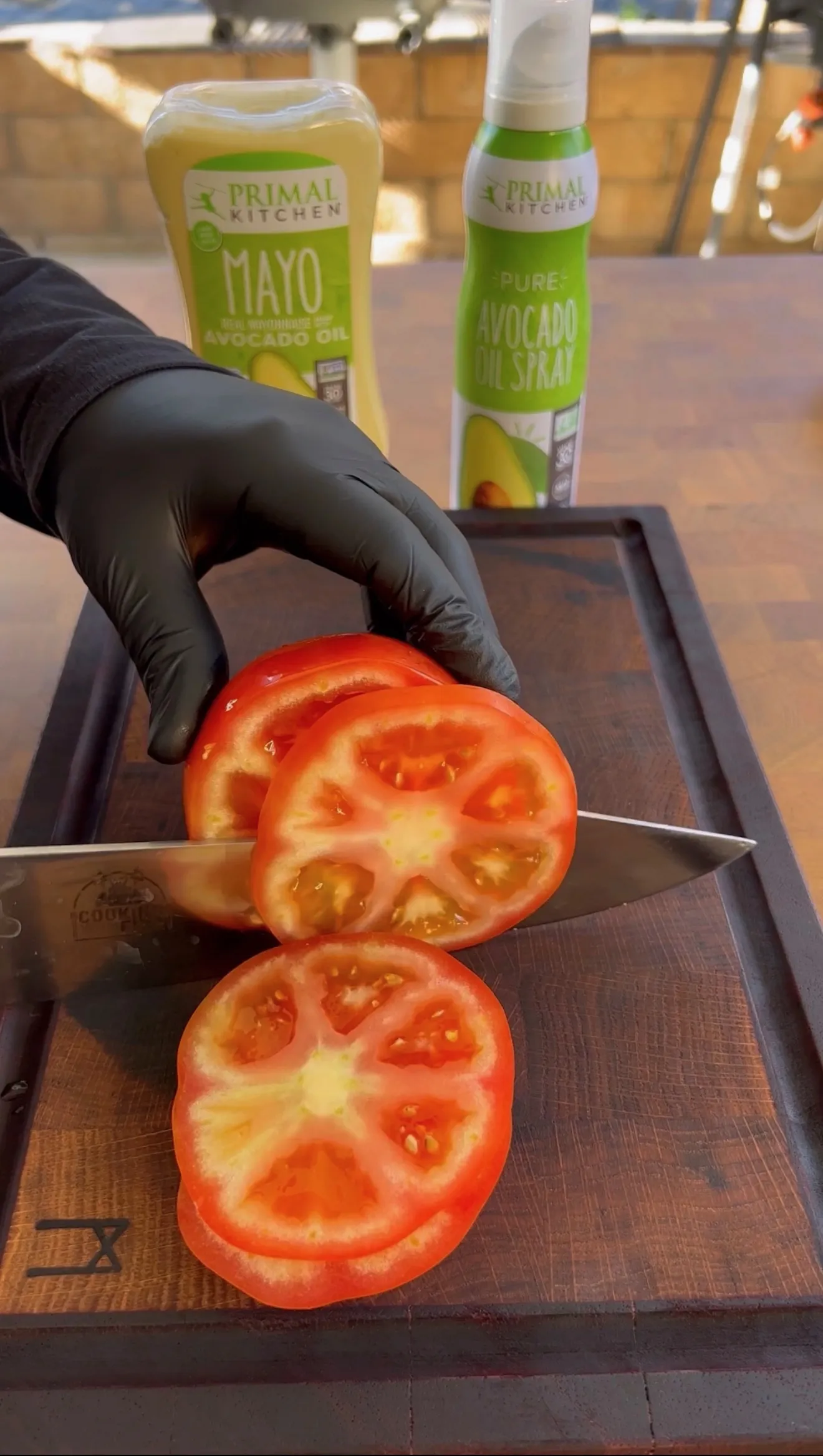 Fresh red tomato being sliced on wooden cutting board with knife and avocado products for BLT sandwich prep