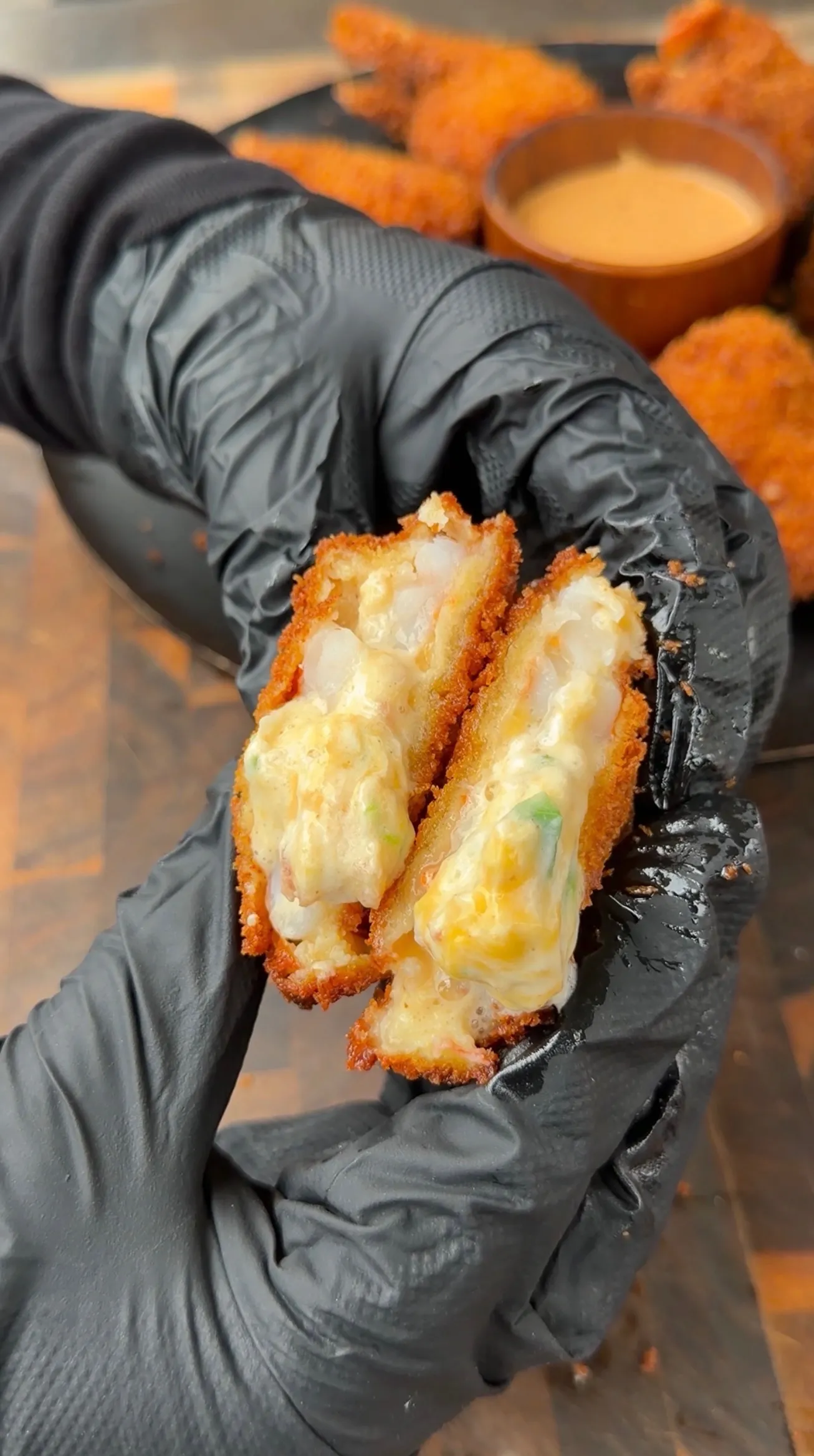 Hands in black gloves holding split fried shrimp stuffed with creamy jalapeño popper filling, showing cheesy interior