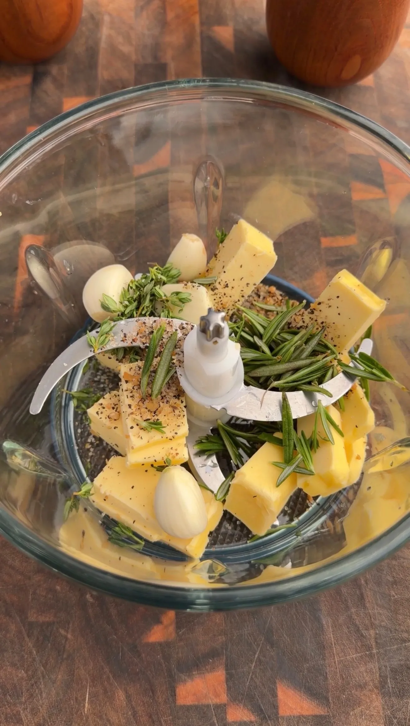 Food processor bowl with cubed butter, fresh garlic cloves, rosemary and thyme herbs with black pepper