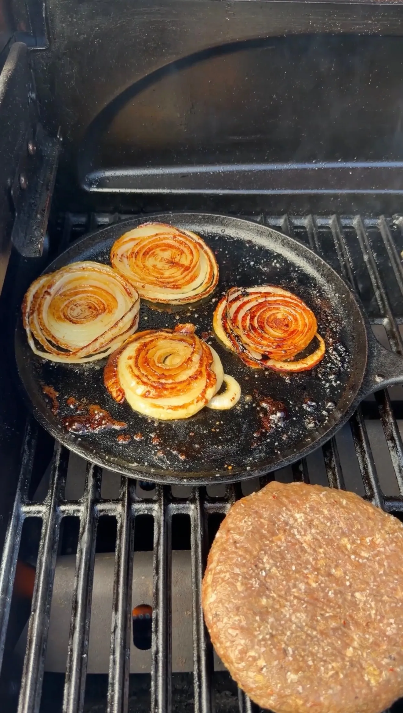 Caramelized onion slices cooking in a cast iron skillet on a gas grill with burger bun visible