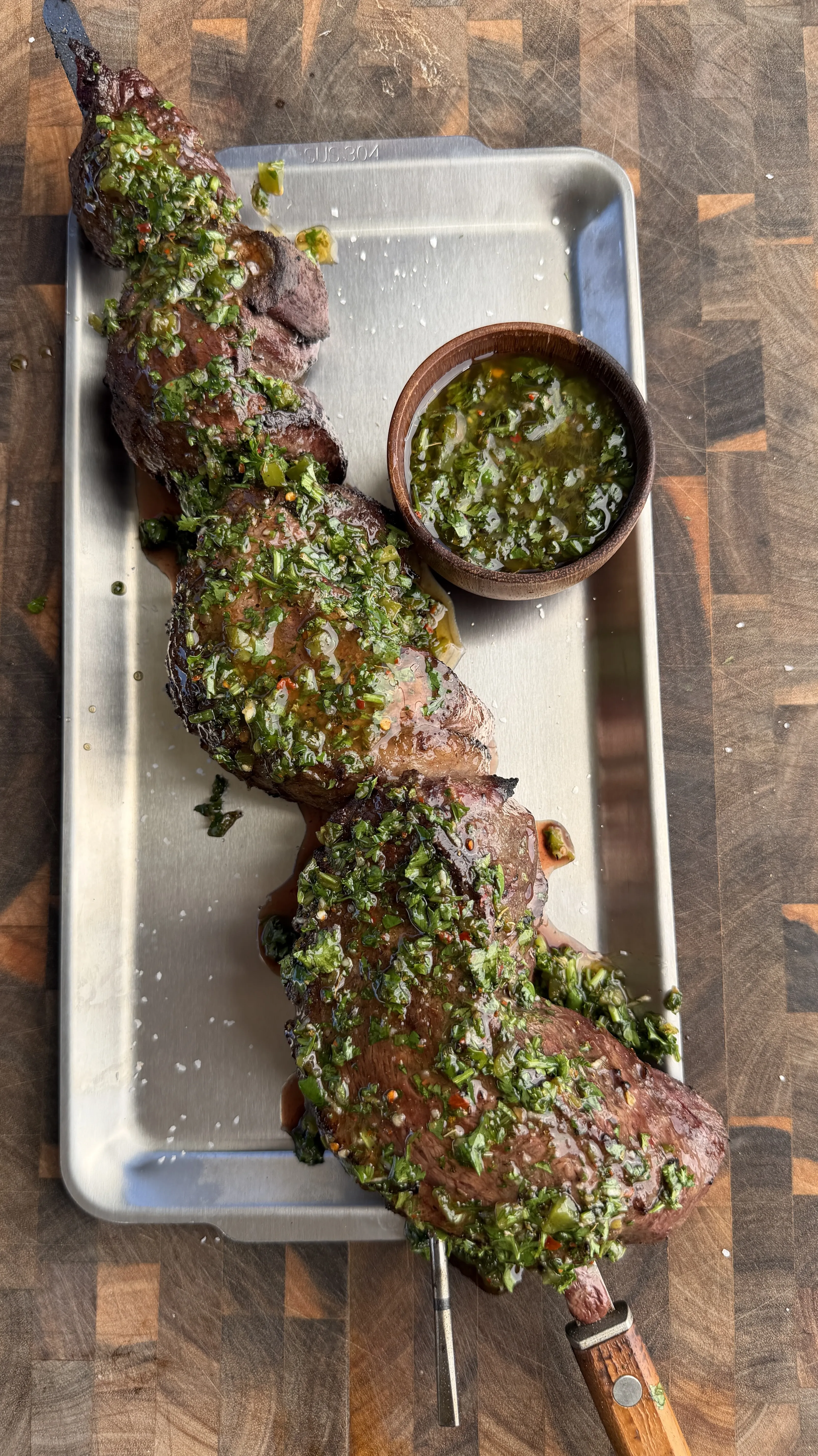 Grilled picanha beef skewer topped with green chimichurri sauce on white tray with sauce bowl