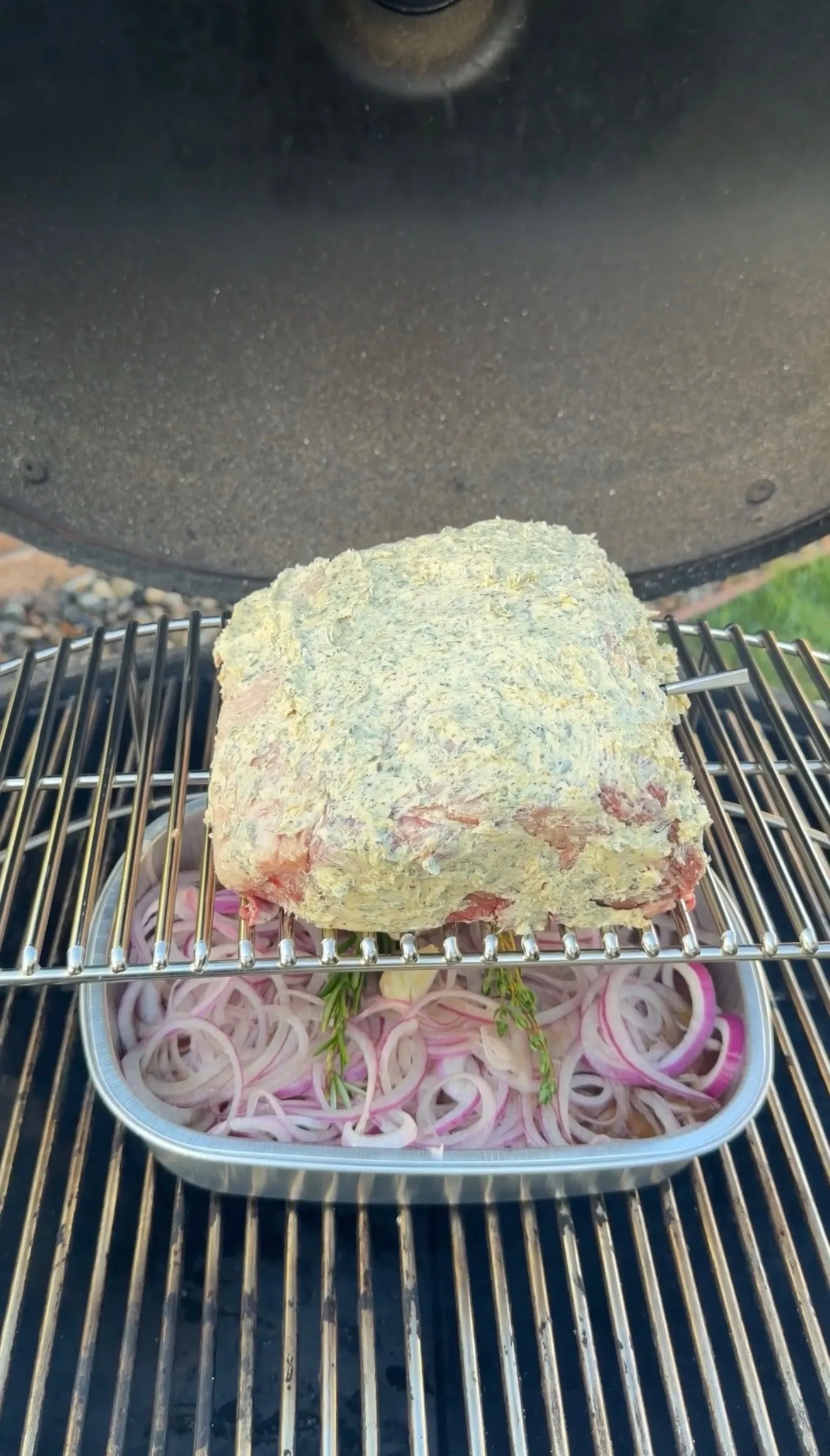 Herb-crusted prime rib roast smoking on grill grates with sliced red onions in aluminum pan underneath