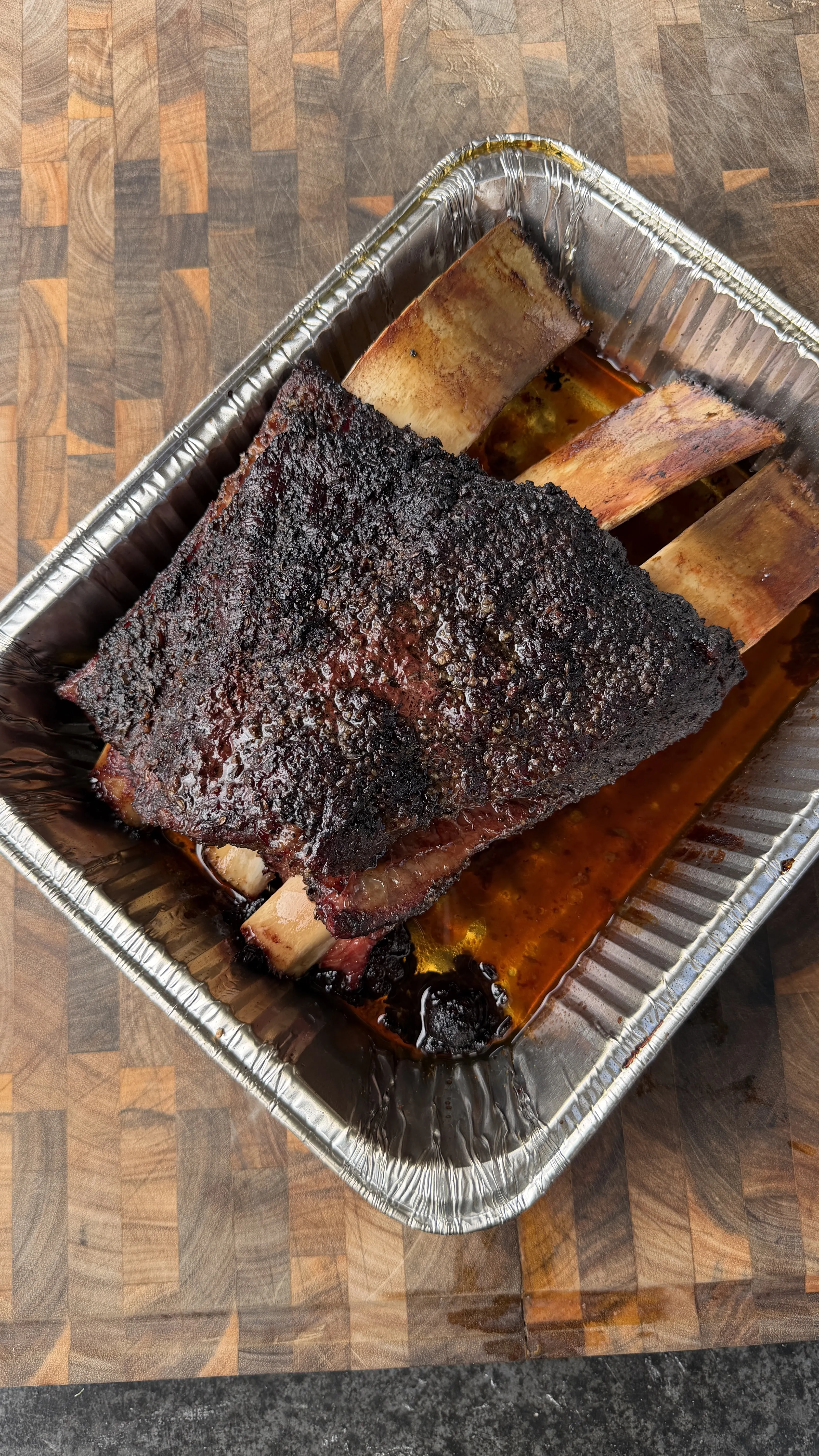 Hickory smoked beef plate ribs with dark bark crust in an aluminum pan on a wood cutting board