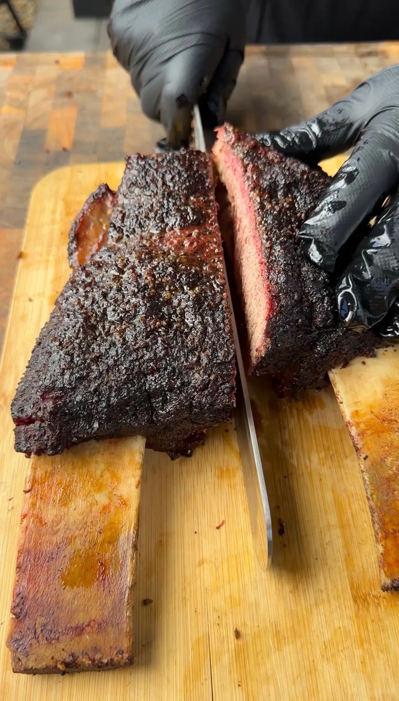 Gloved hands slicing hickory smoked beef plate ribs with dark bark and pink smoke ring on wooden cutting board