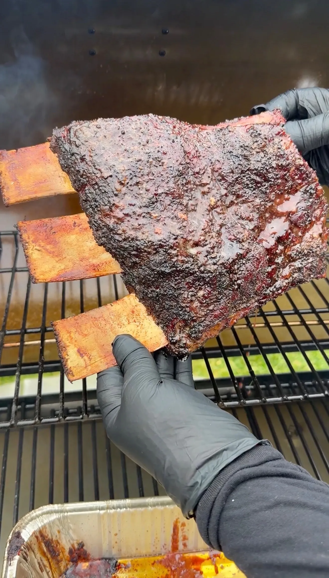 Hands in black gloves lifting a large smoked beef plate rib with dark bark crust from a BBQ smoker grill