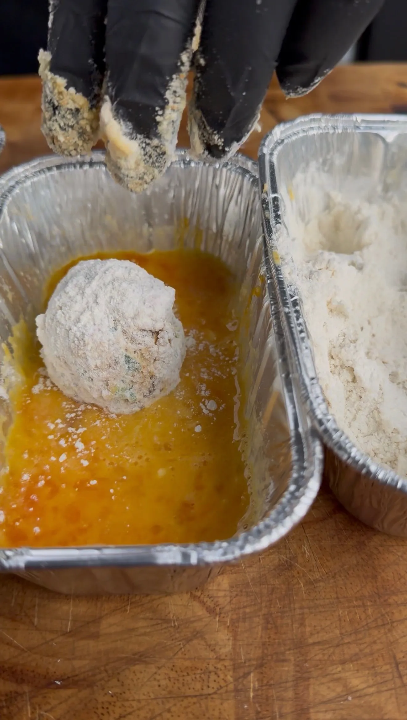 Flour-coated jalapeño popper bite being dipped in egg wash in aluminum tray next to flour breading station