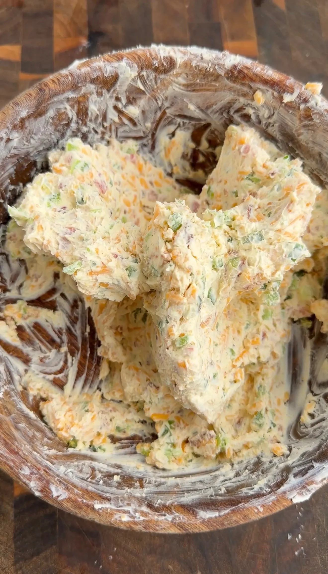 Cream cheese filling with shredded cheddar, jalapeños, and bacon bits in a wooden mixing bowl