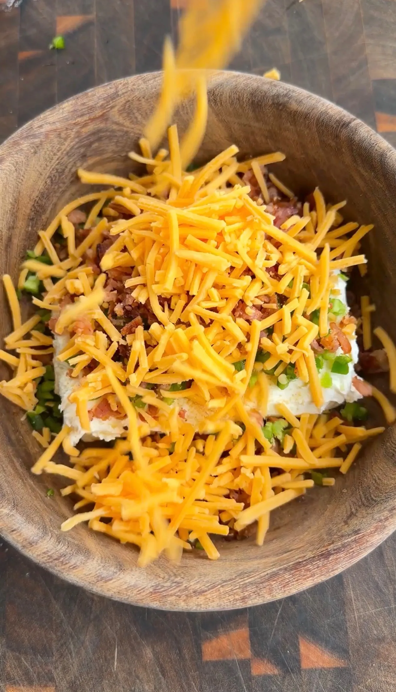 Shredded cheddar cheese being added to jalapeño popper stuffing with cream cheese and bacon in a wooden bowl