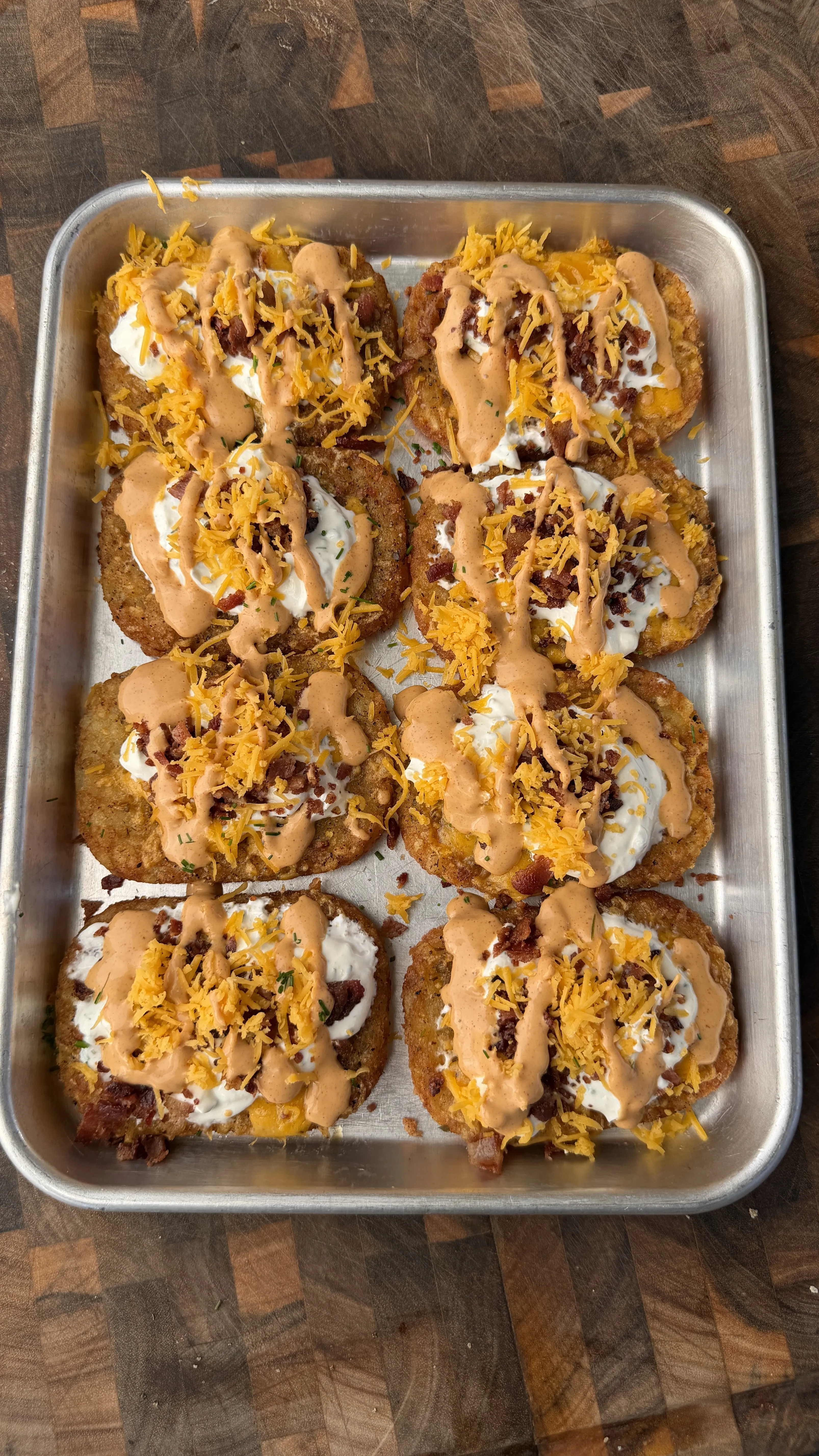 Eight loaded crispy hash brown patties topped with sour cream, cheddar cheese, bacon bits, and sauce on baking sheet
