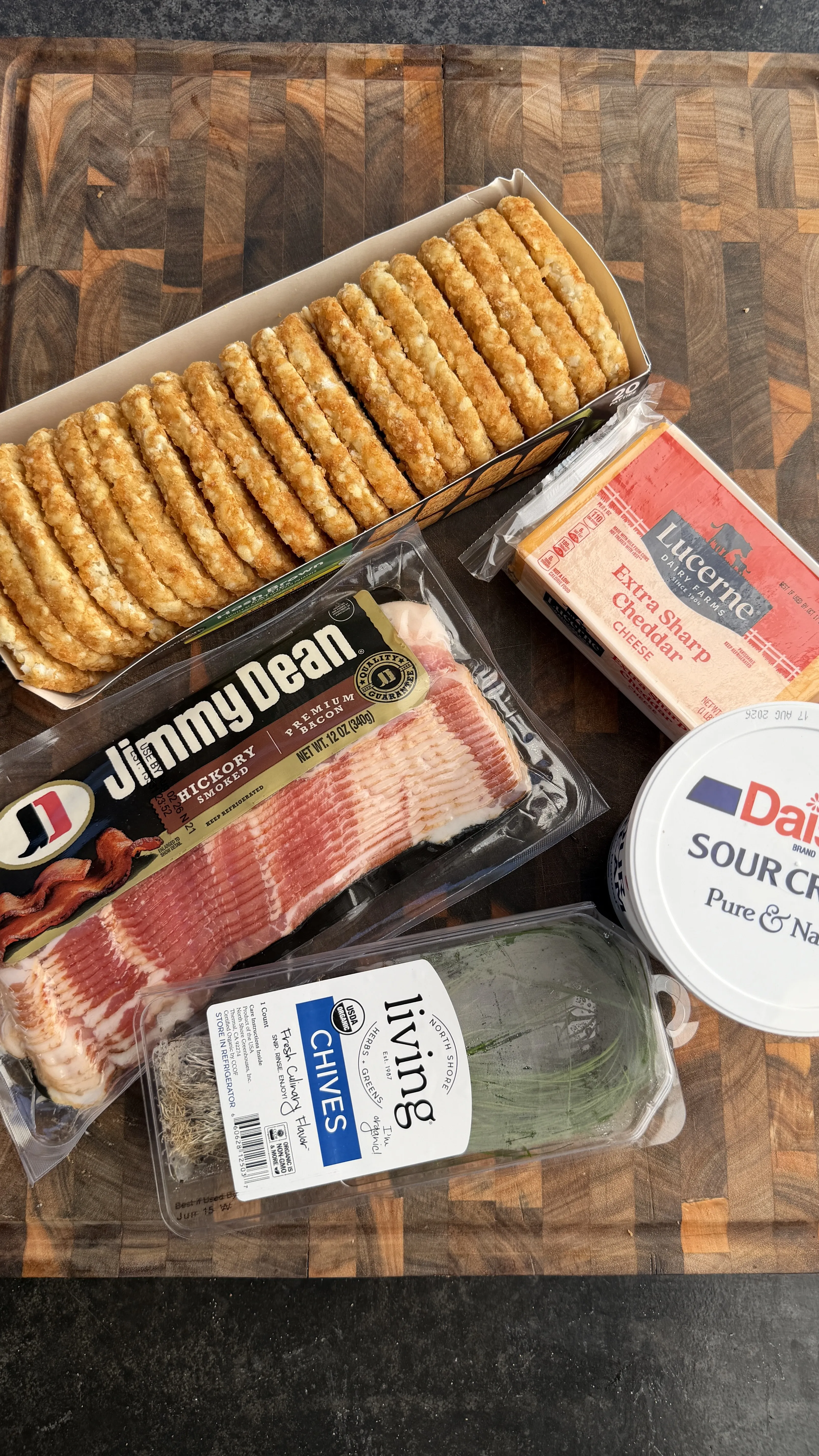 Ingredients for loaded hash browns including bacon, cheese, sour cream, chives and crackers on wooden cutting board
