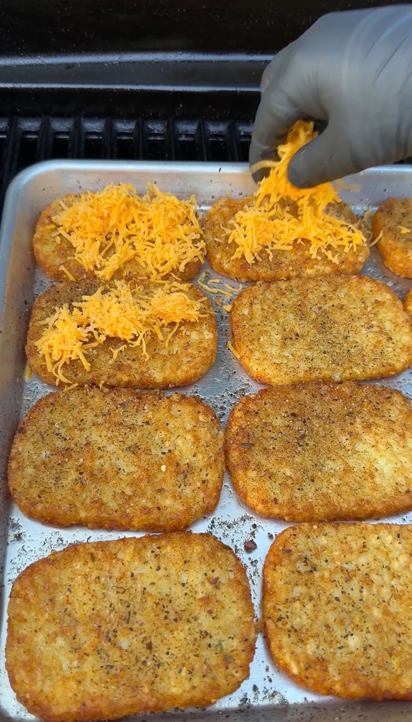 Golden crispy hash brown patties on baking sheet being topped with shredded cheddar cheese by hand