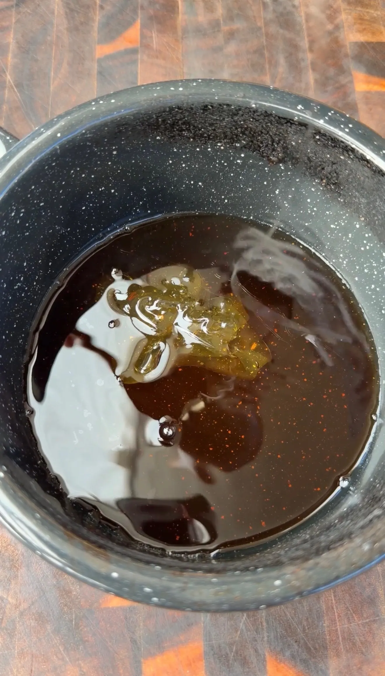 Dark maple bourbon glaze simmering in a speckled black saucepan on a wooden surface