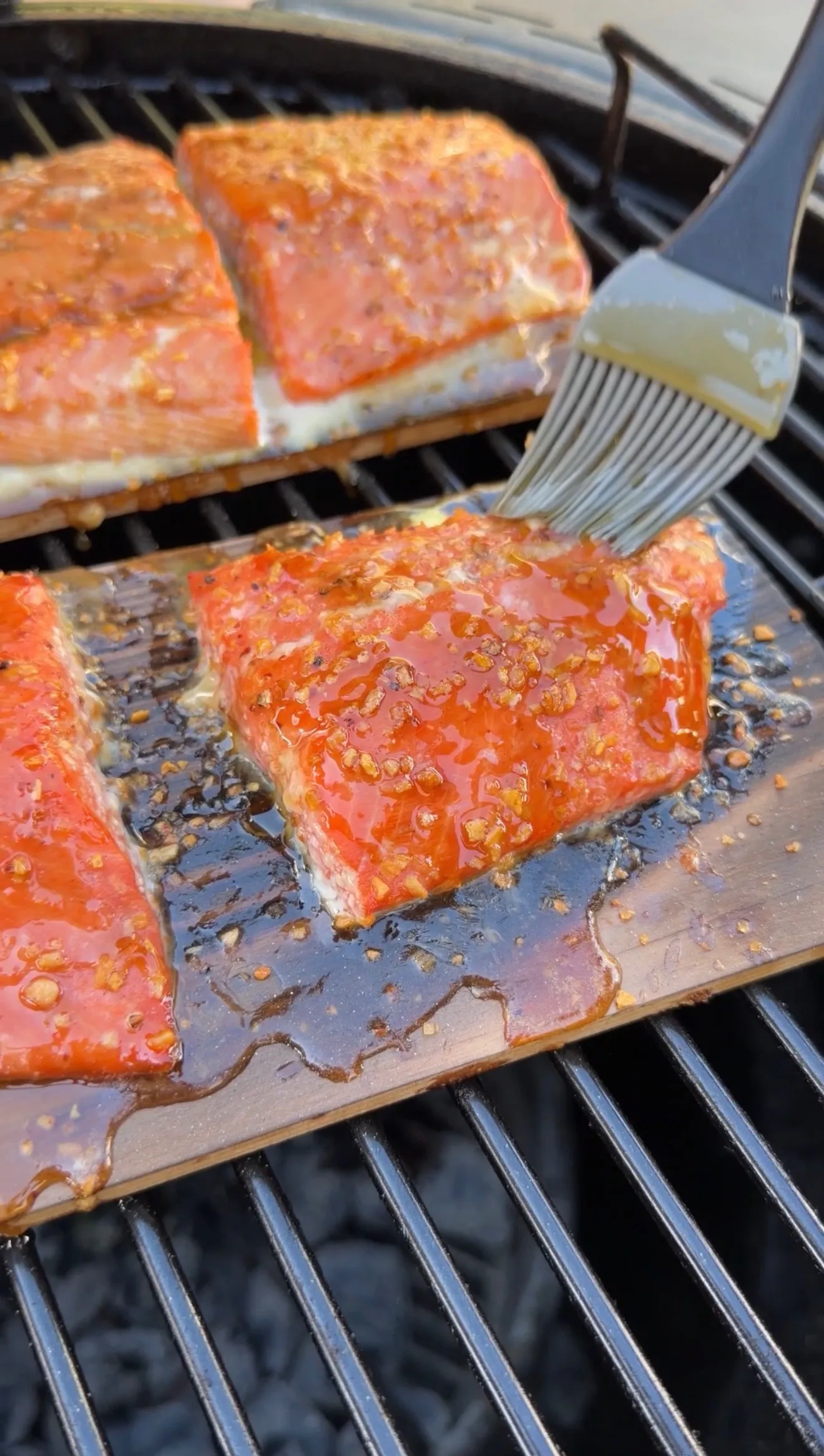 Glazed maple bourbon salmon fillets on cedar planks being basted with sauce on an outdoor grill