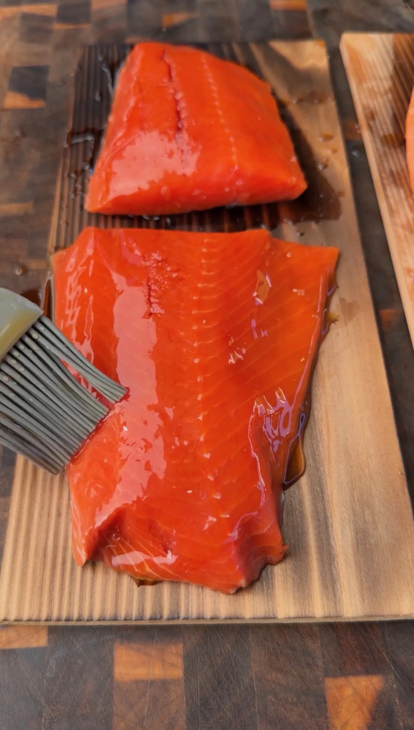 Raw salmon fillets being brushed with maple bourbon glaze on a cedar plank before smoking