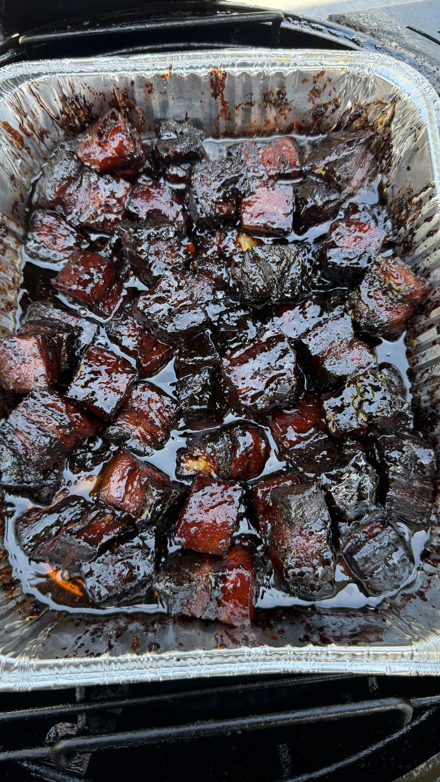 Glossy molasses-cured smoked pork belly burnt ends in aluminum foil pan with caramelized edges