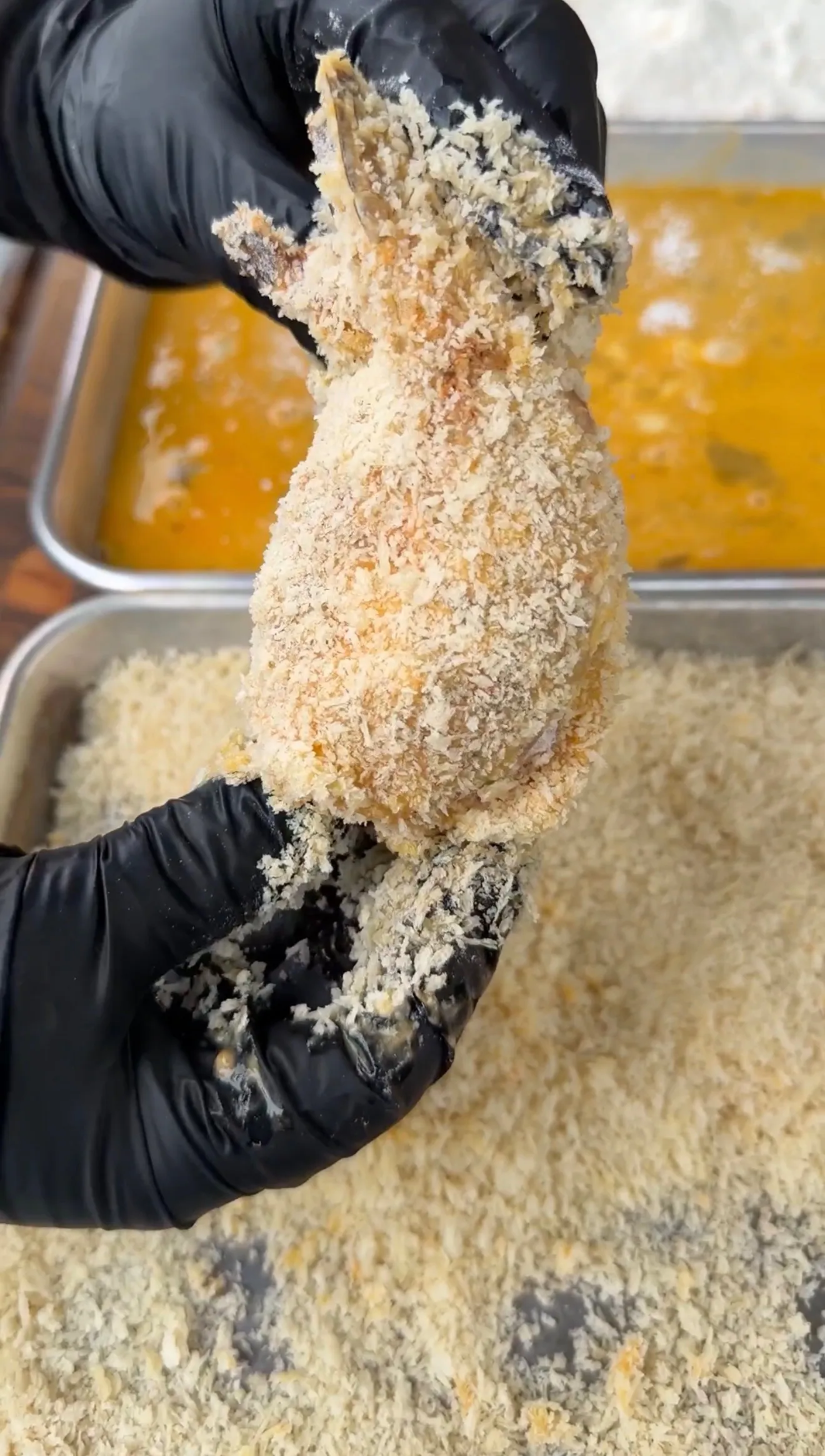 Hands in black gloves coating a jalapeño popper stuffed shrimp in panko breadcrumbs with egg wash tray behind