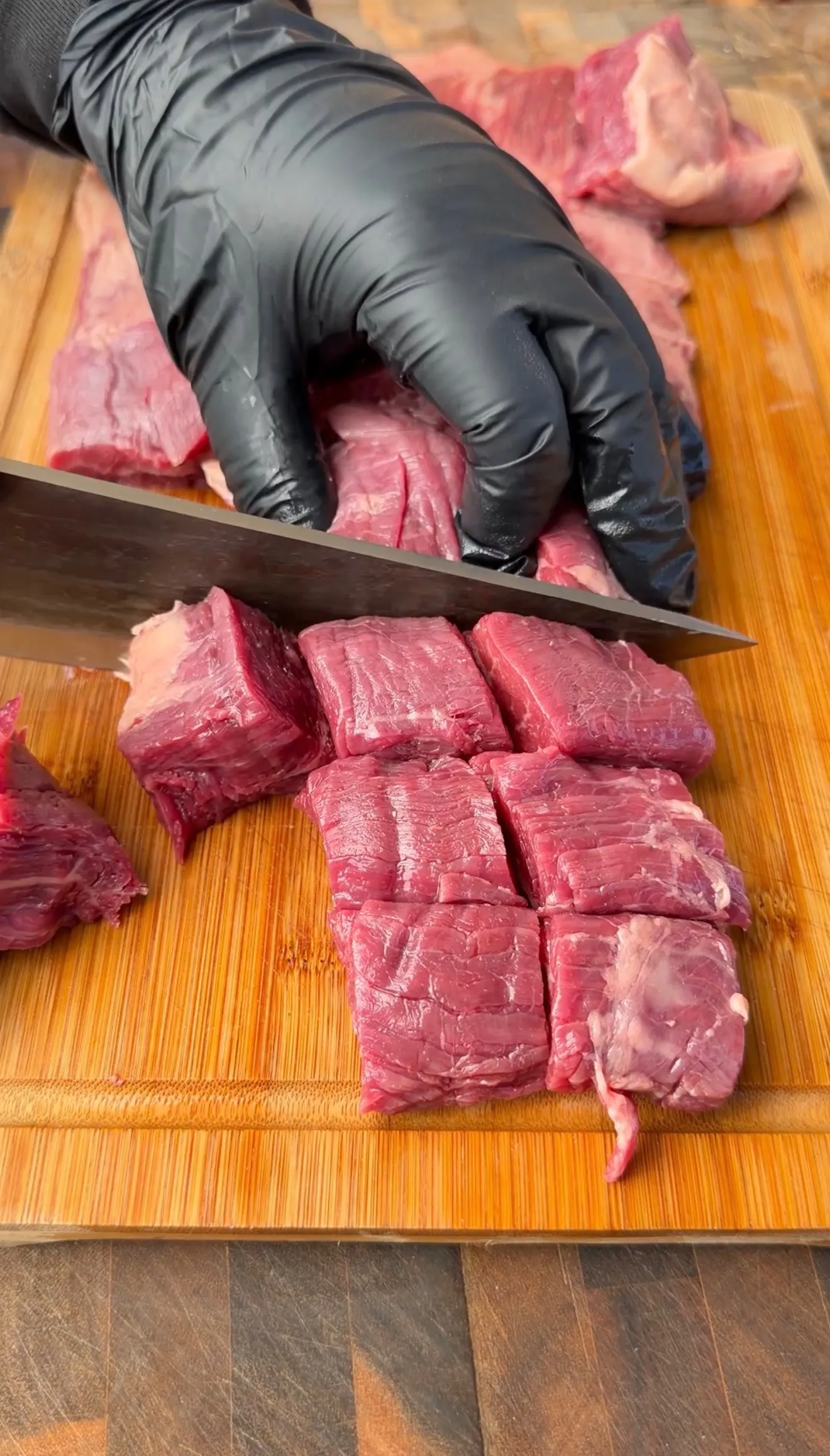 Fresh raw beef being cut into cubes on wooden cutting board with knife for teriyaki skewers preparation