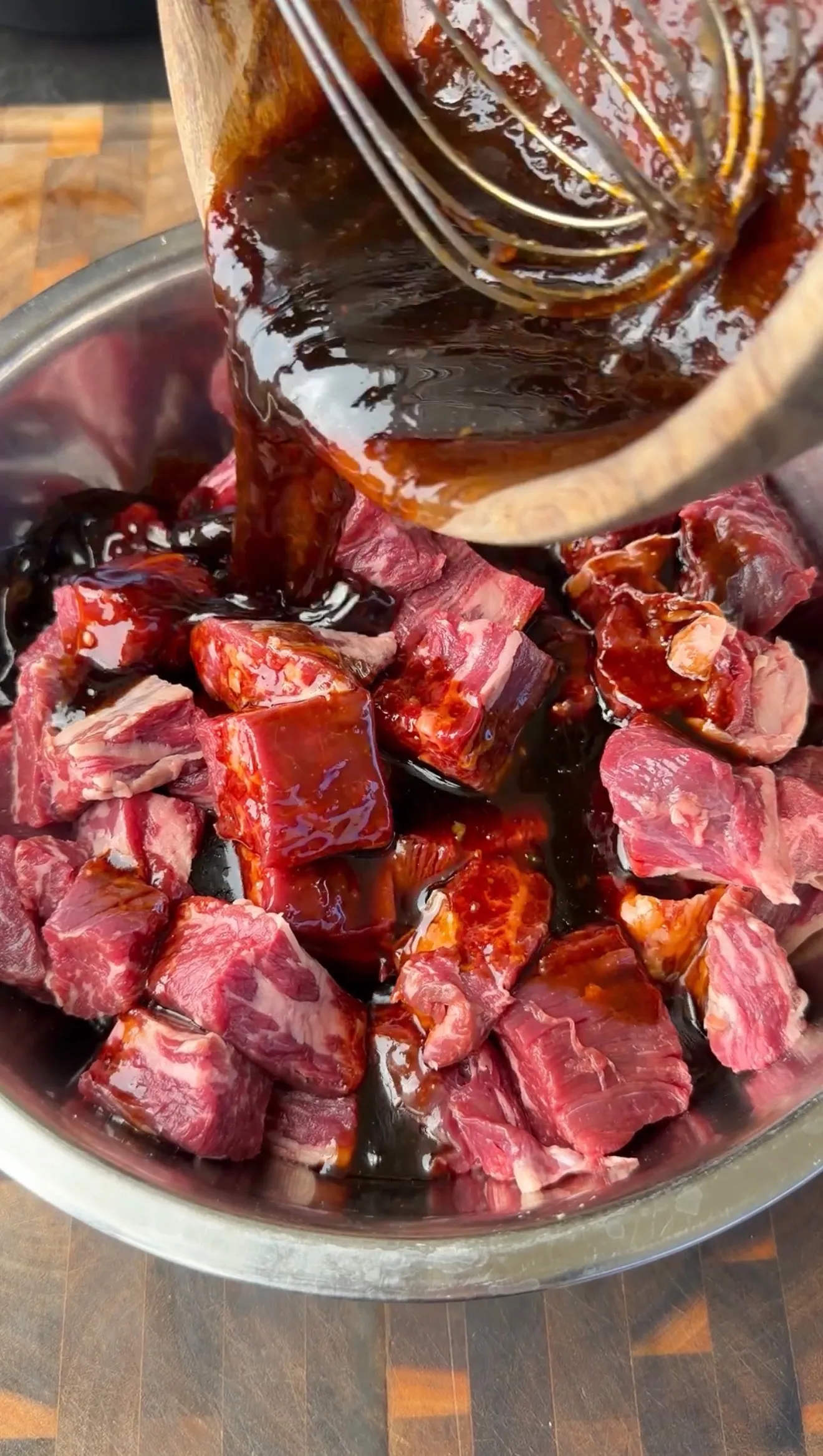 Raw beef cubes marinating in dark teriyaki sauce being poured from wooden spoon in metal bowl