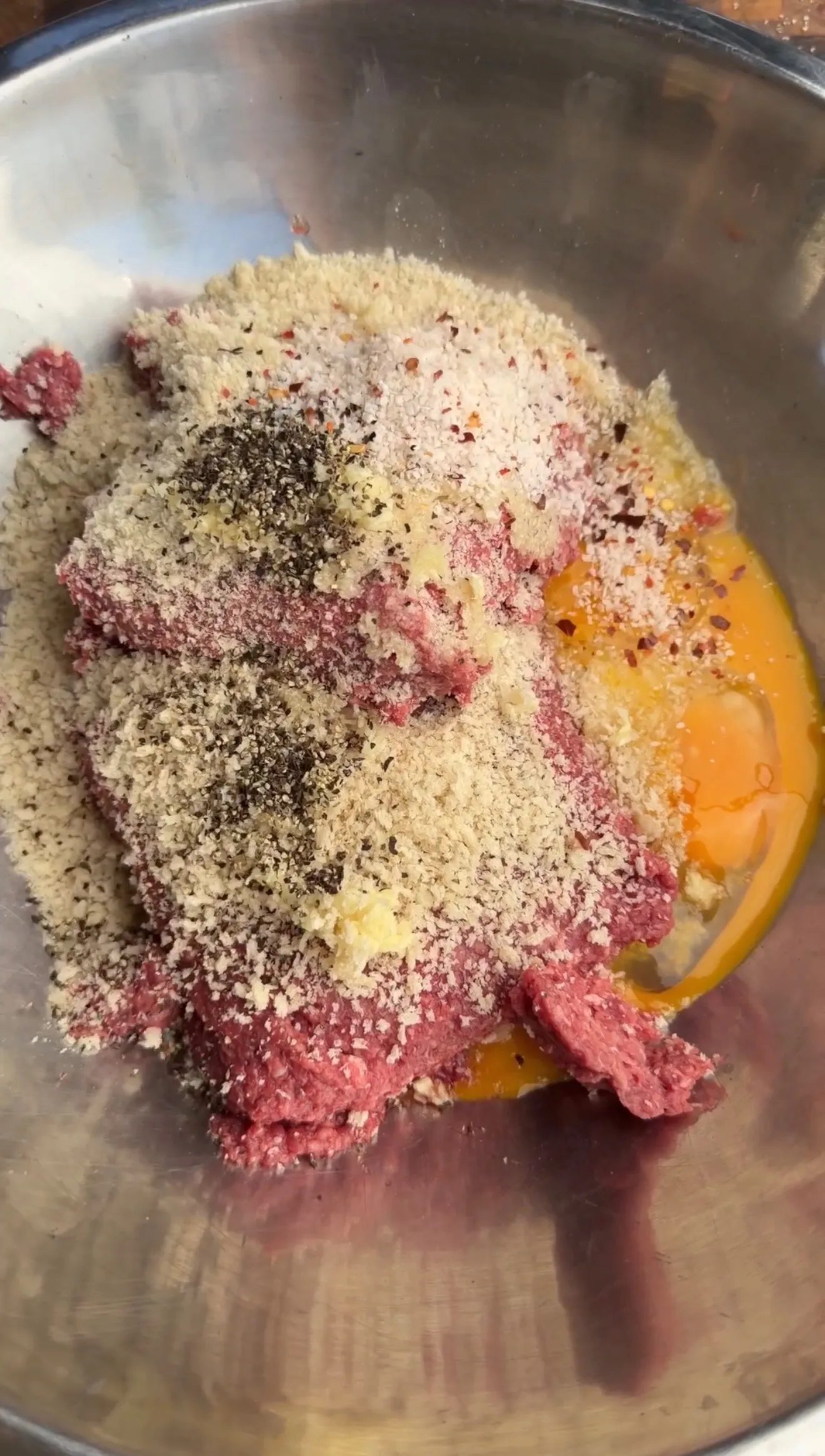 Raw ground bison meat mixed with breadcrumbs, eggs, and seasonings in a stainless steel mixing bowl