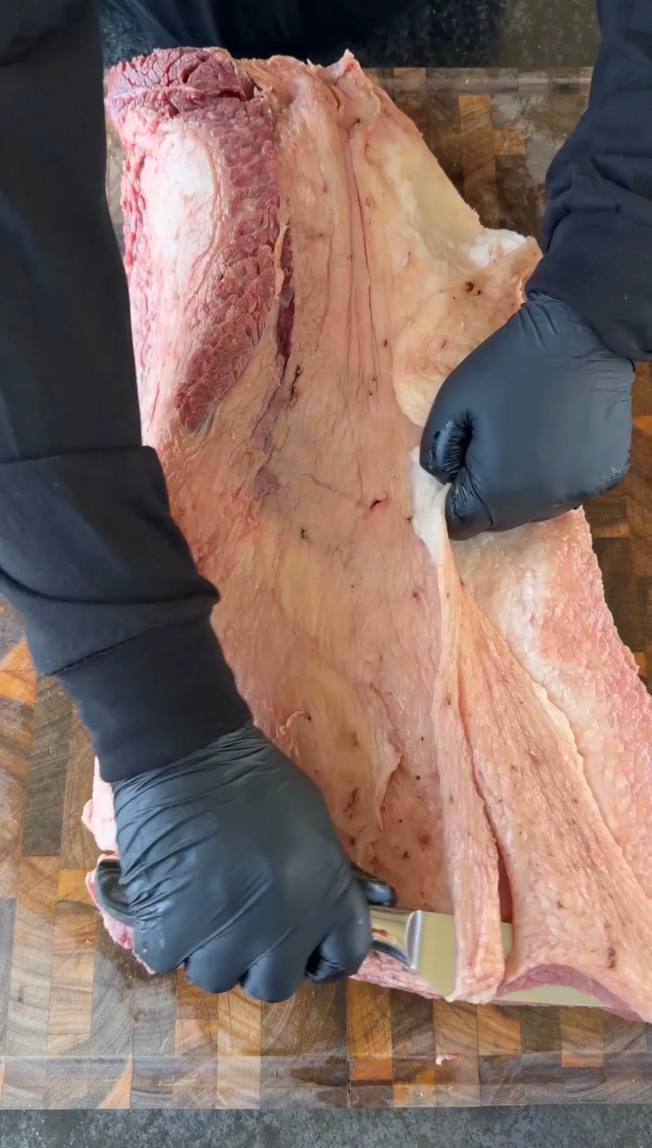 Hands in black gloves trimming excess fat from a large raw beef brisket on a wooden cutting board