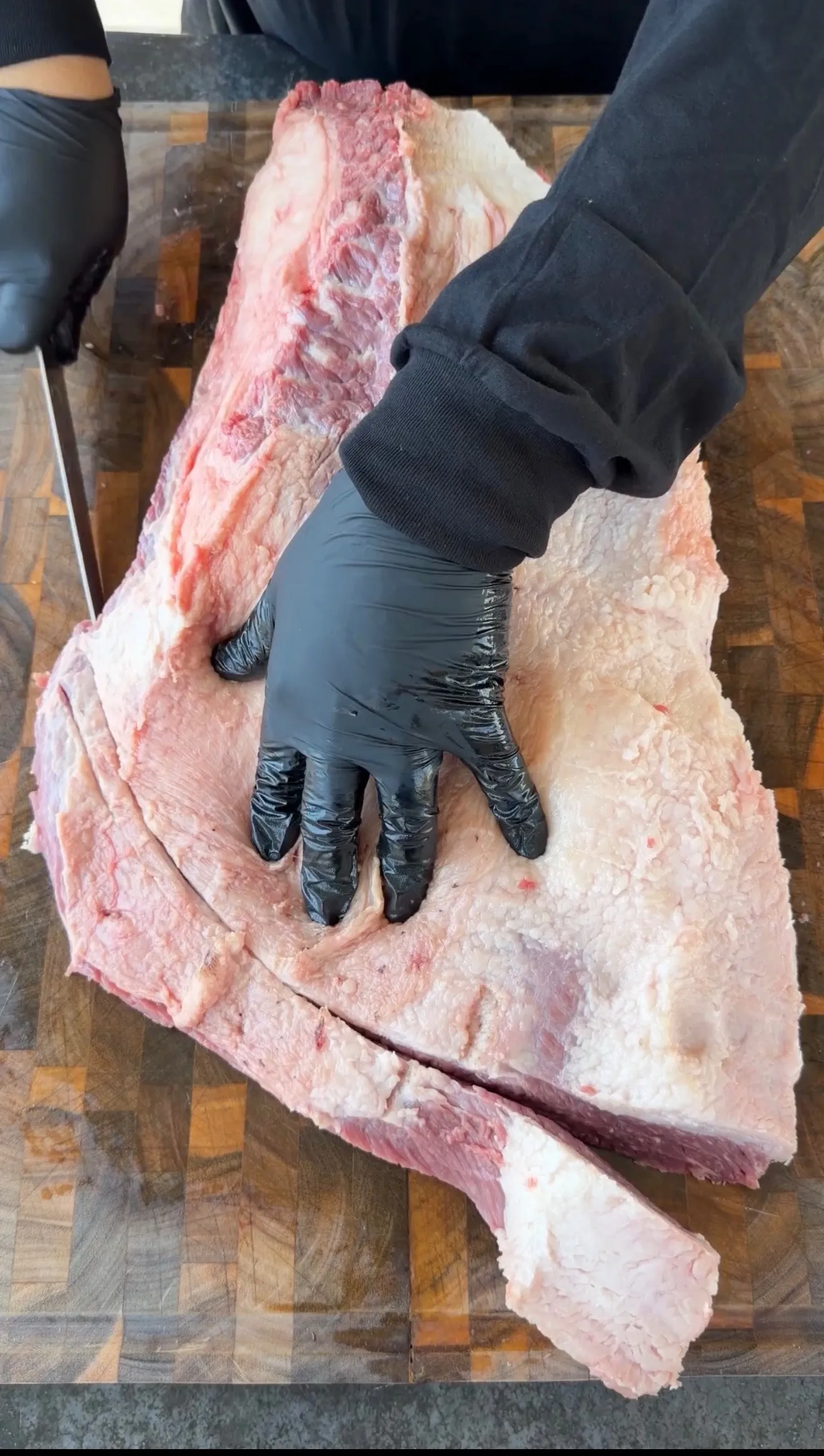 Gloved hands trimming a large raw beef brisket on a wooden butcher block cutting board