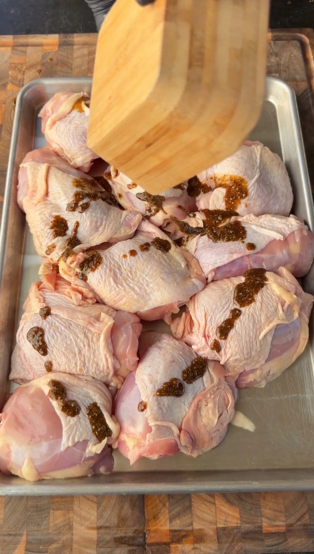 Raw chicken thighs on a sheet pan being coated with Cajun lemon pepper honey marinade using a wooden spatula