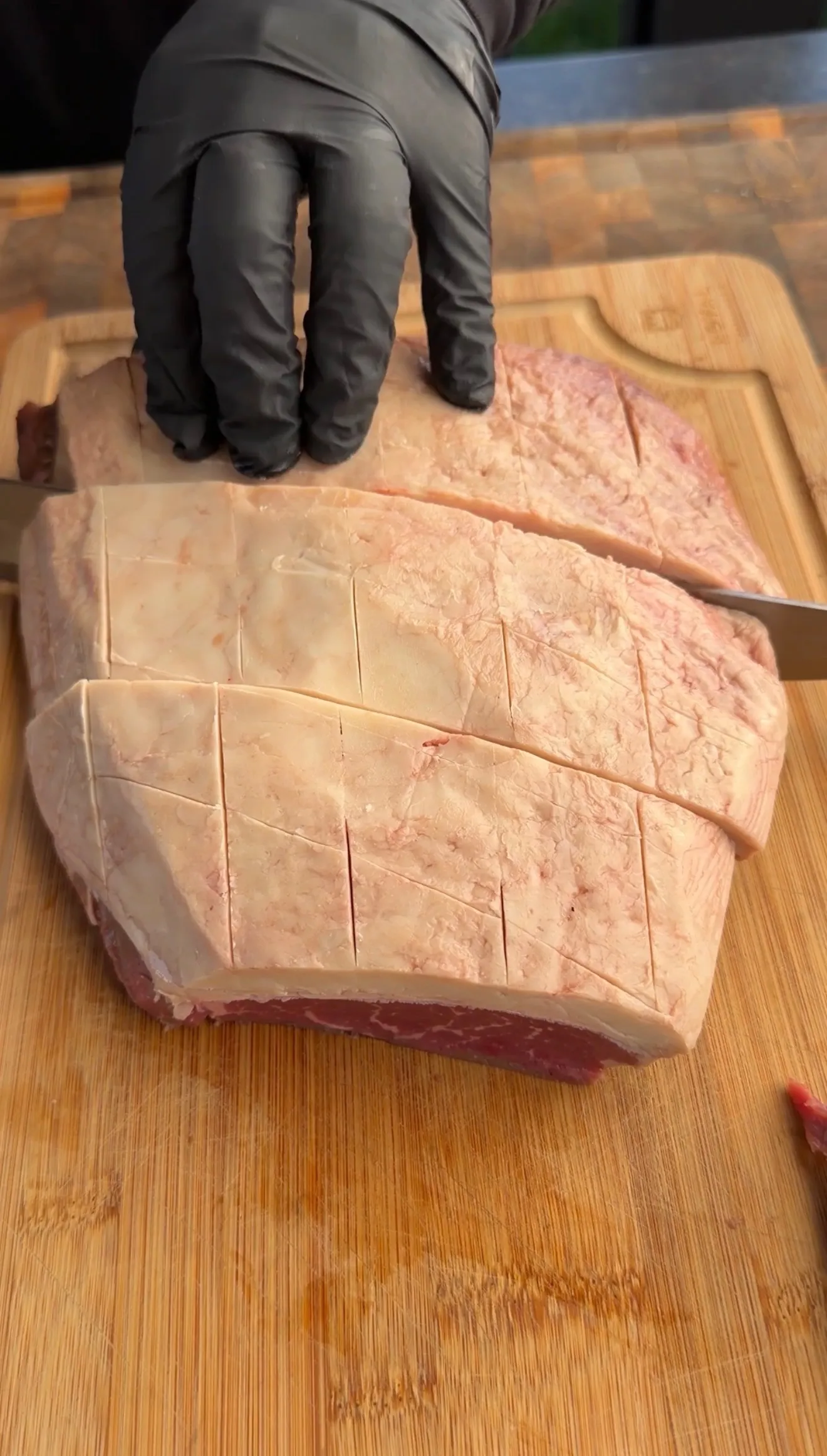 Raw picanha steak with crosshatch scoring marks on wooden cutting board, hands in black gloves using knife