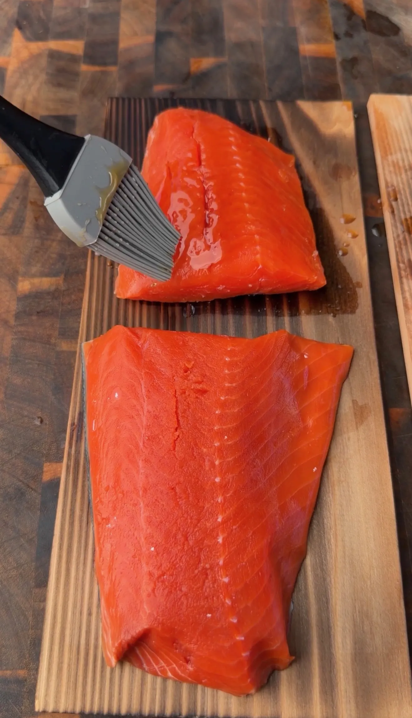 Two raw salmon fillets being brushed with maple bourbon glaze on cedar planks before smoking
