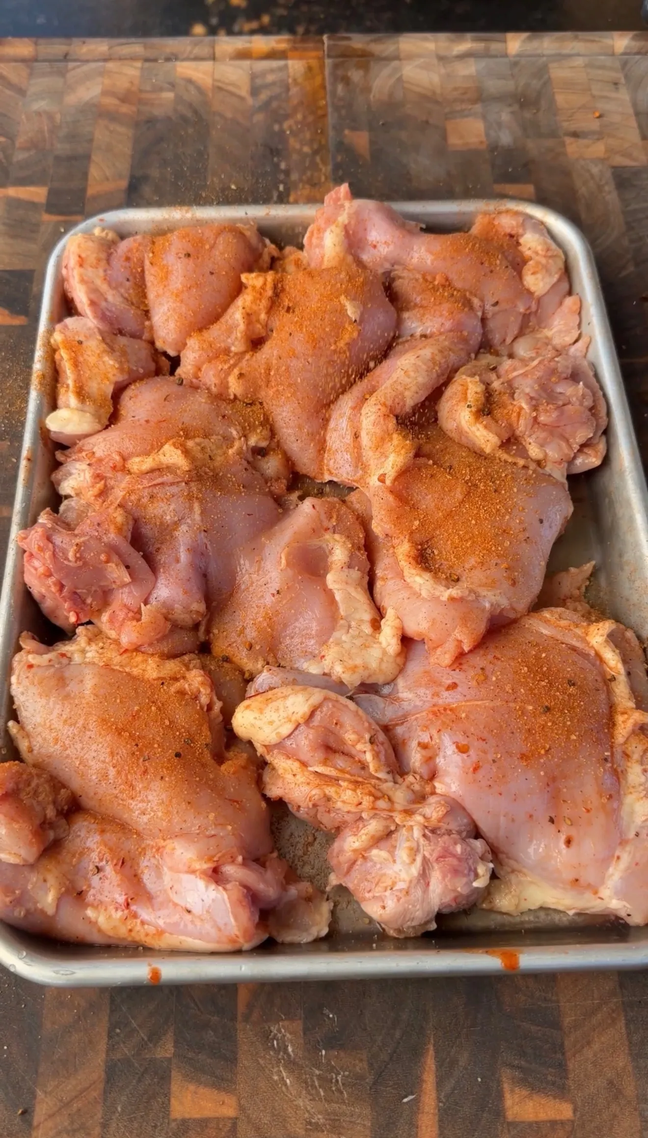 Raw boneless chicken thighs seasoned with a spicy red rub in a metal baking pan on a wooden cutting board