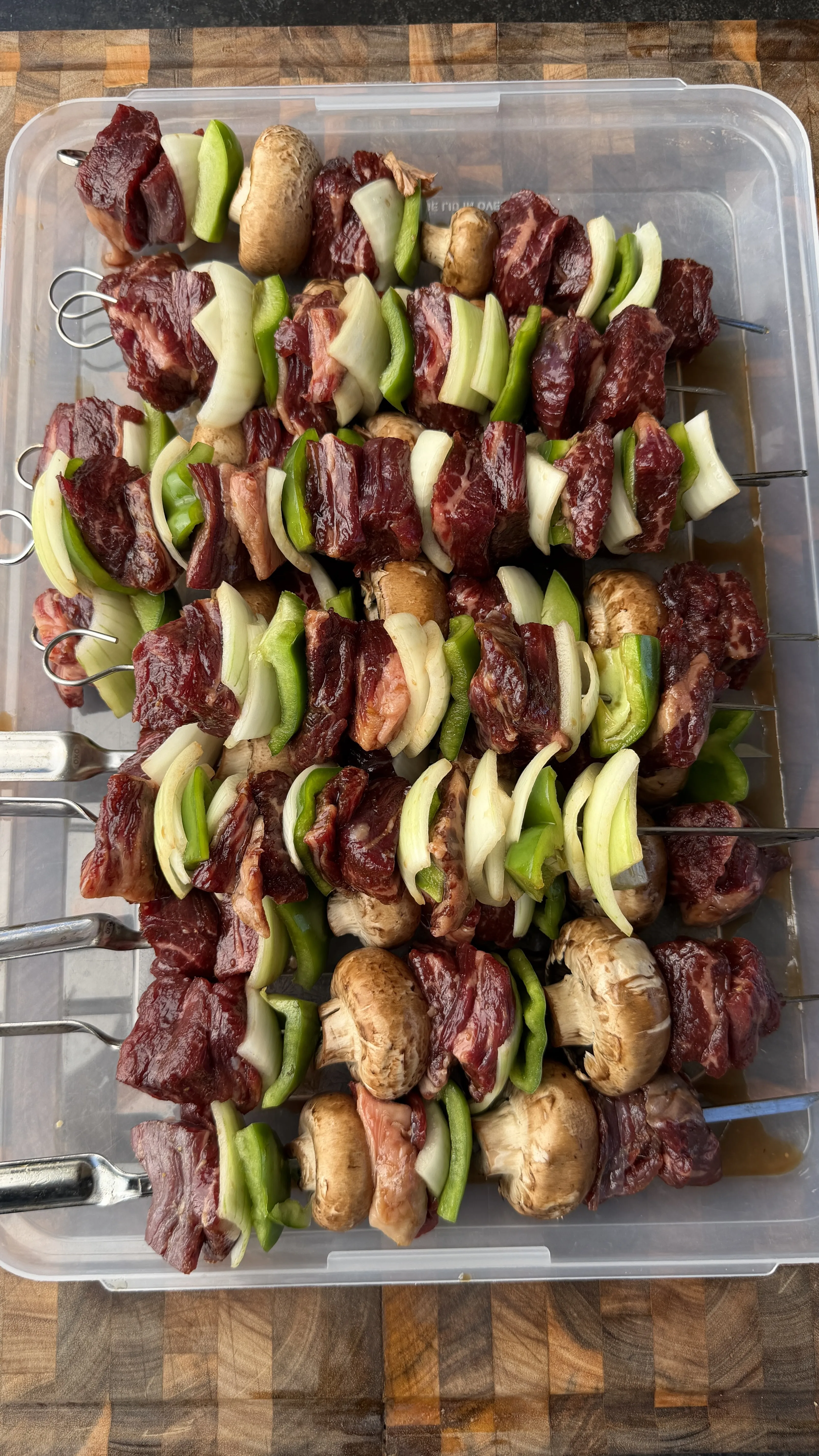 Raw teriyaki beef skewers with mushrooms, bell peppers and onions in clear container ready for grilling