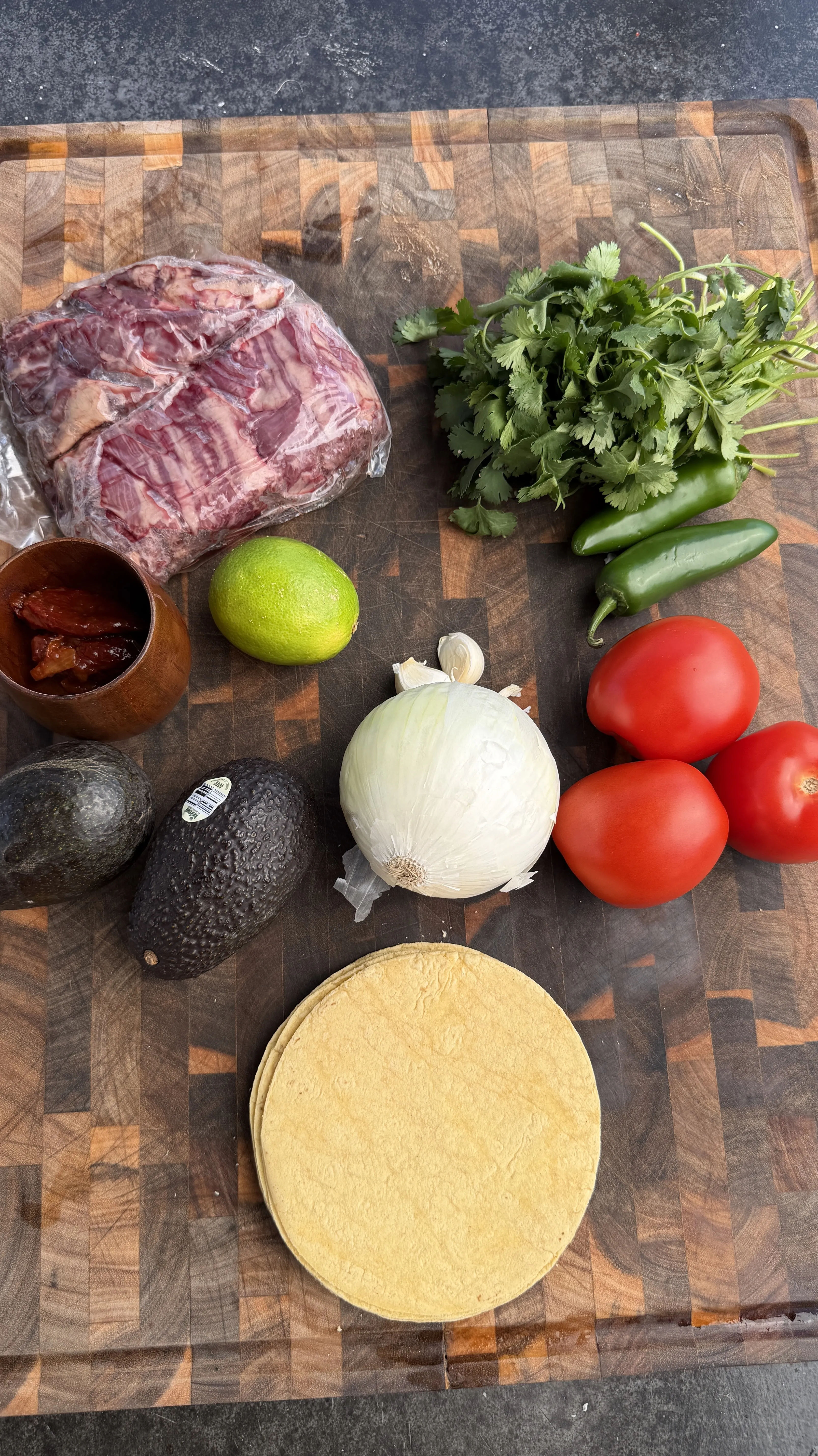 Raw skirt steak and fresh taco ingredients including tortillas, tomatoes, avocados, cilantro and lime on wooden board