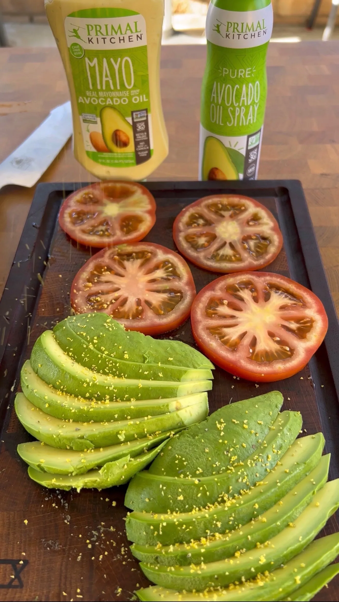 Sliced avocado and tomato arranged on dark cutting board with Primal Kitchen mayo and avocado oil spray