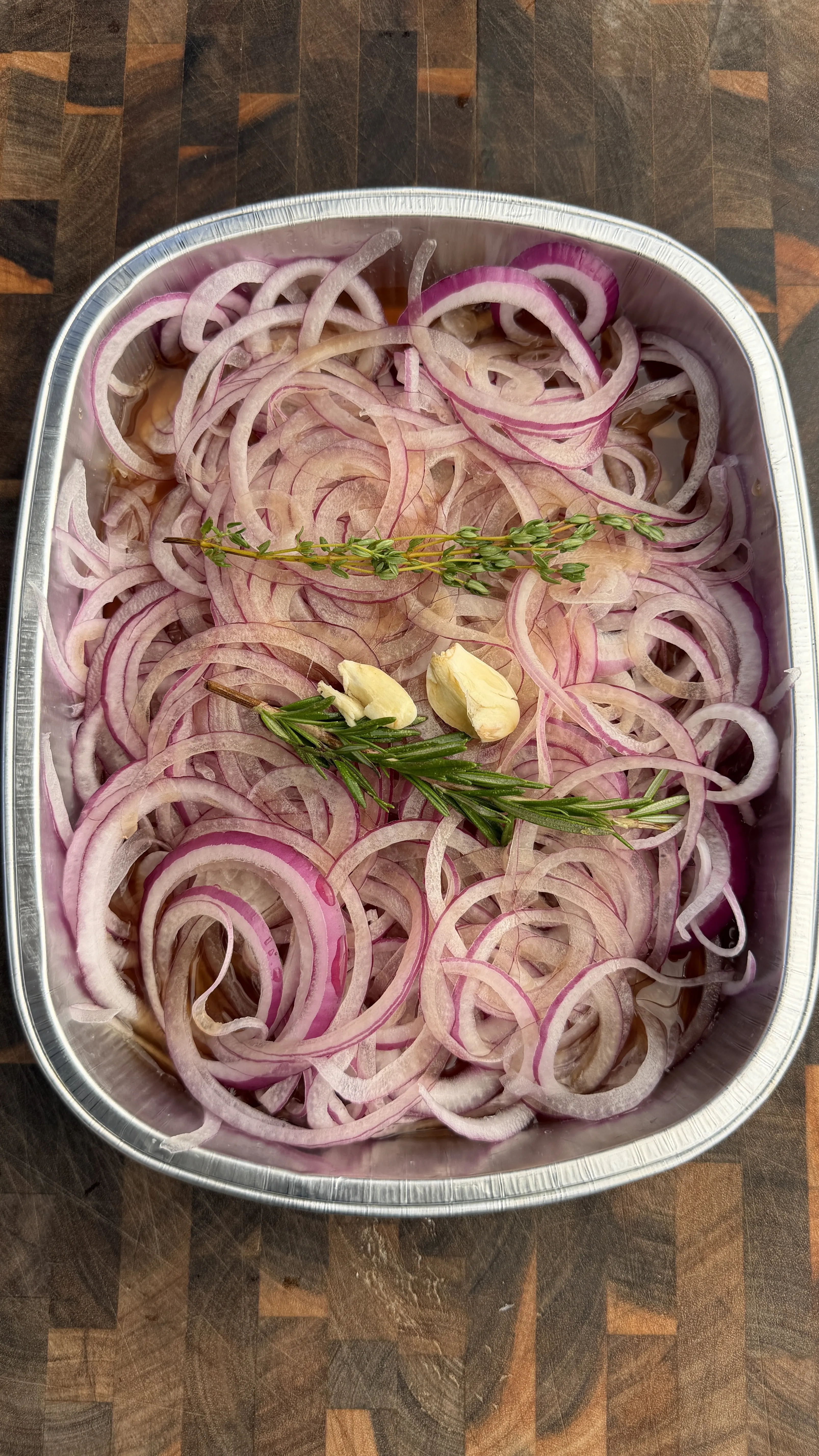 Thinly sliced red onions with fresh herbs and garlic cloves in aluminum baking pan for prime rib sandwich prep