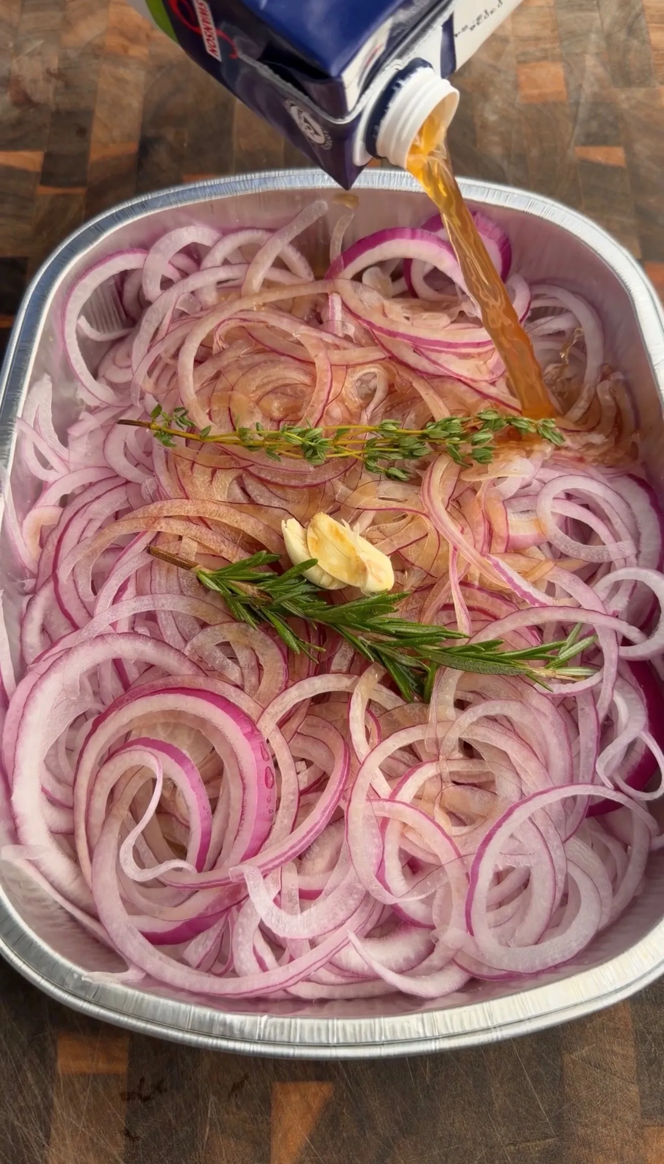 Thinly sliced red onions with fresh herbs and garlic in aluminum pan being drizzled with oil