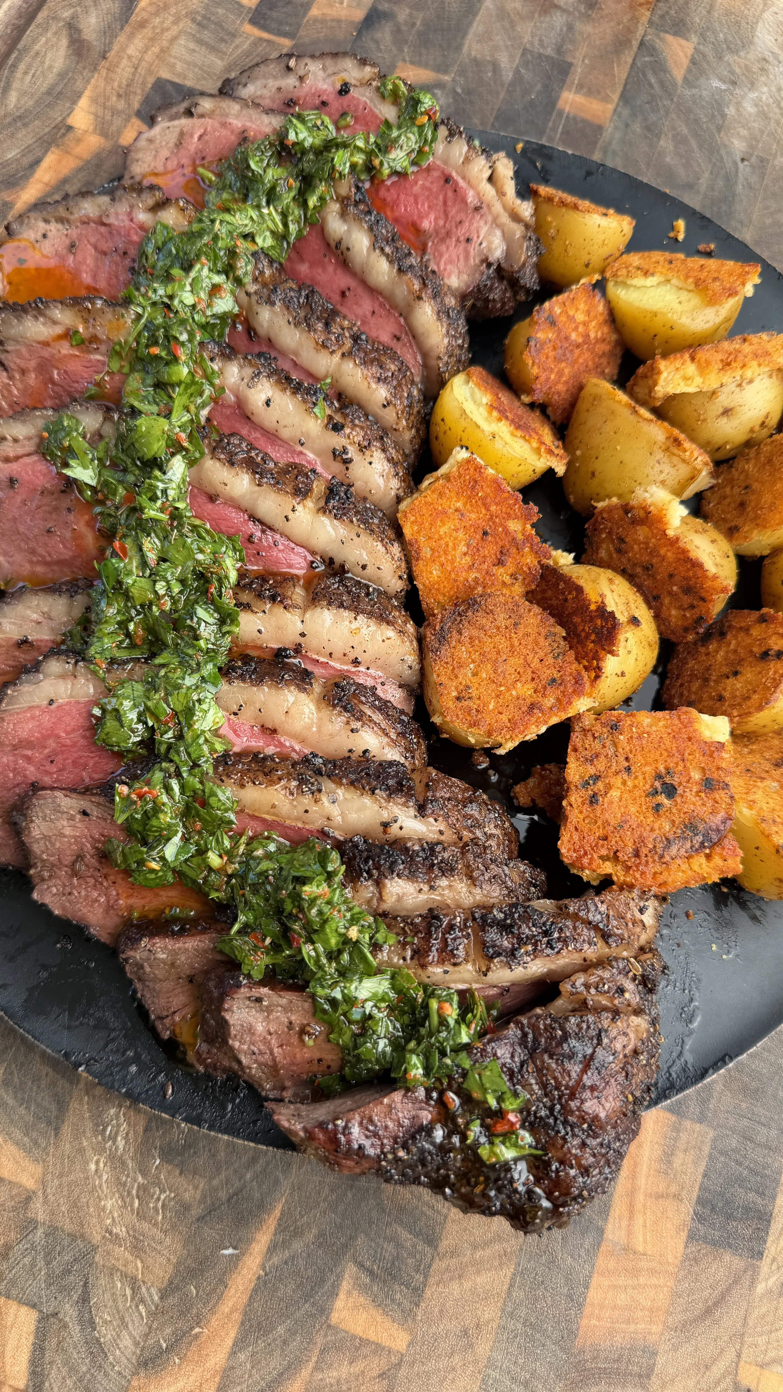 Sliced smoked picanha roast with green chimichurri sauce and golden crispy parmesan potatoes on slate plate