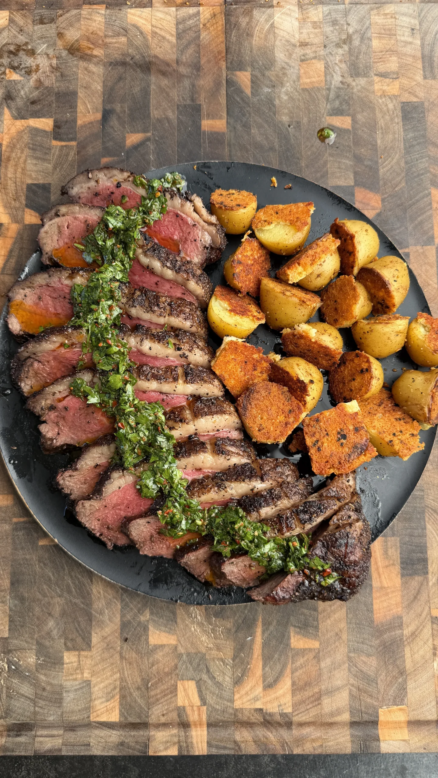 Sliced smoked picanha roast with green chimichurri sauce and golden crispy Parmesan potatoes on dark plate