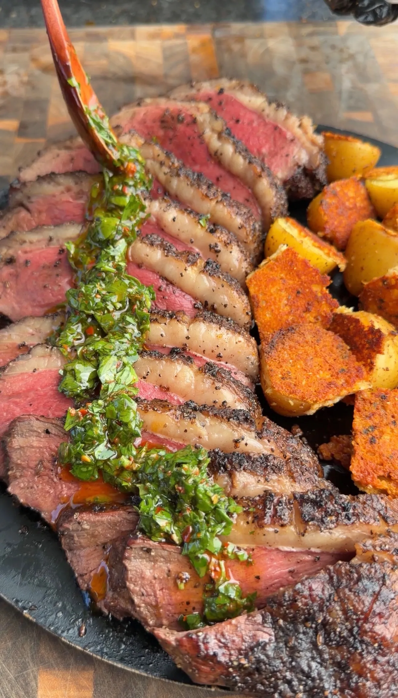 Sliced smoked picanha roast with green chimichurri sauce and crispy parmesan potatoes on a dark slate plate