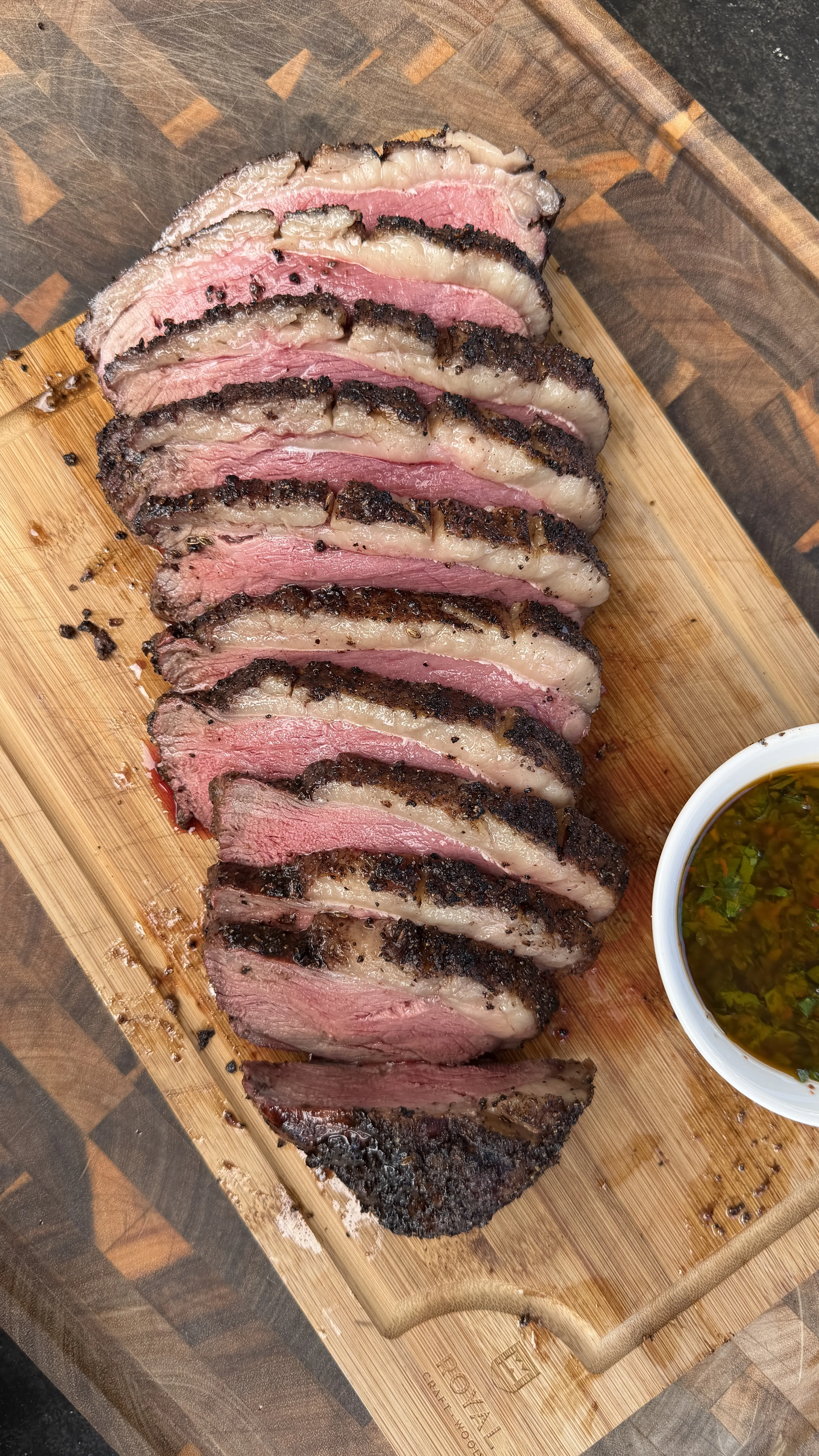 Sliced smoked picanha roast showing pink interior and dark crust on wooden board with green chimichurri sauce