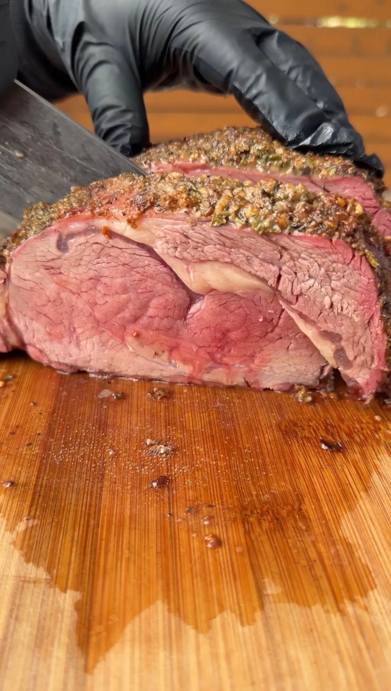 Sliced smoked prime rib with herb crust showing pink interior being cut on wooden cutting board with gloved hands