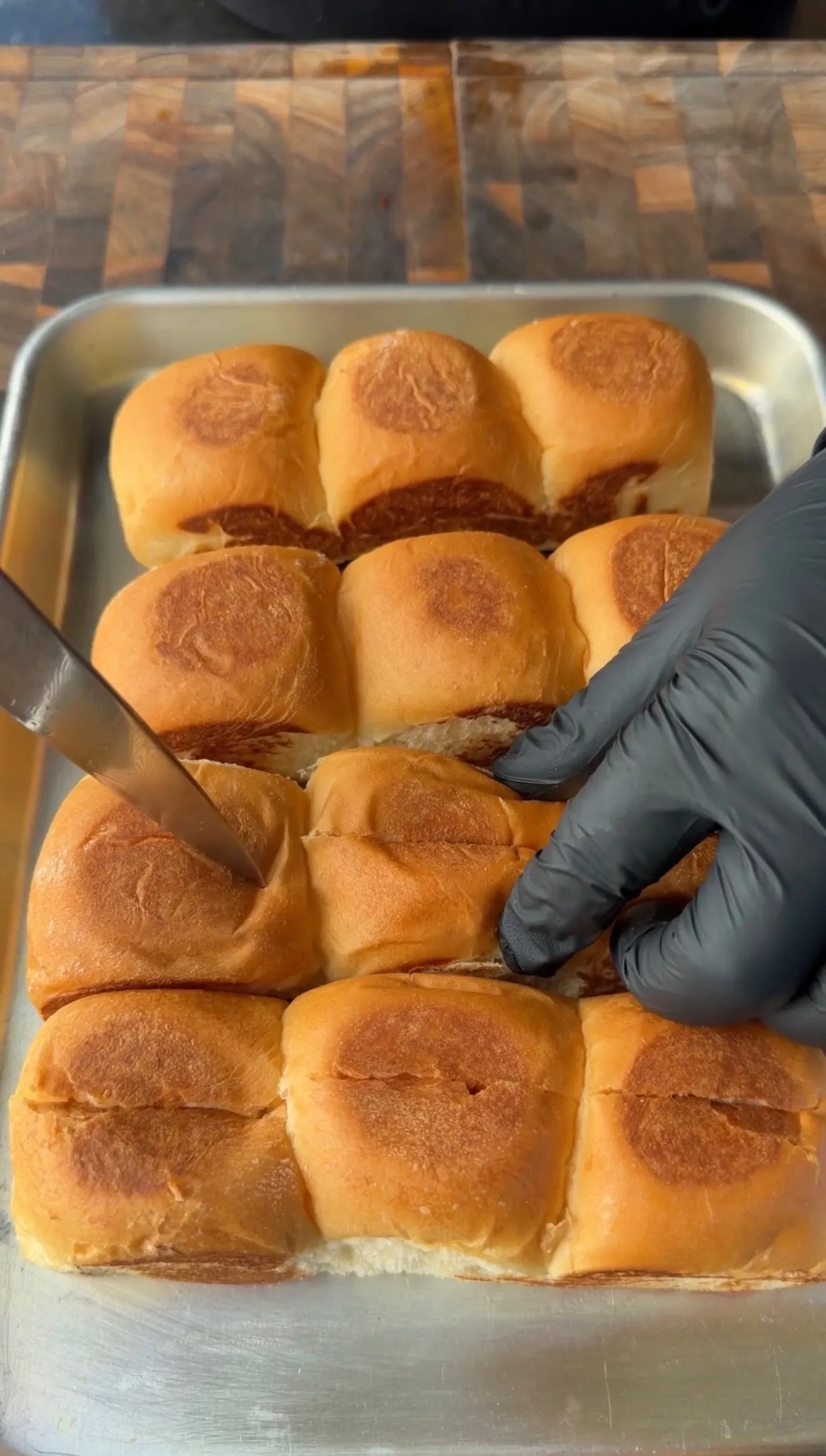Gloved hand slicing Hawaiian rolls horizontally in a sheet pan to prepare Avocado BLT Chopped Sliders