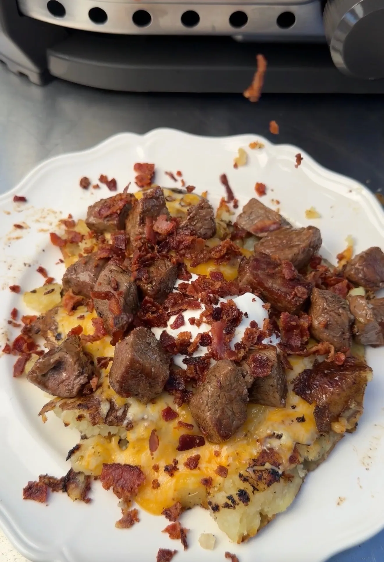 Smashed loaded potatoes topped with steak bites, crispy bacon, melted cheddar, and sour cream on a white plate