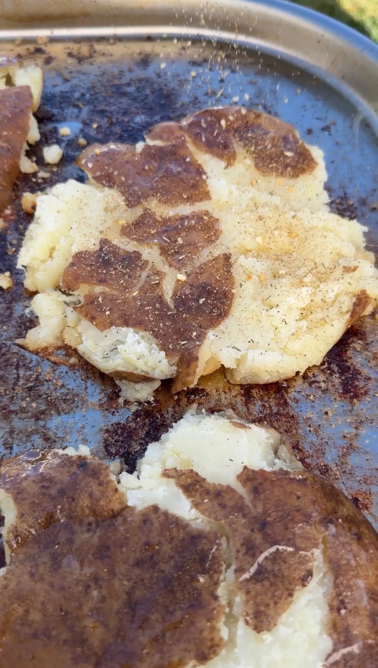 Smashed loaded potato with crispy golden skin being seasoned with spices on a dark baking sheet