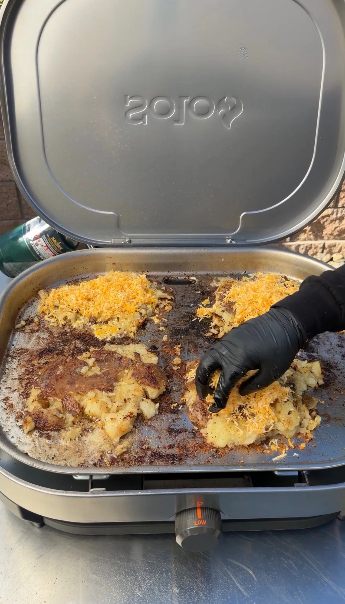 Smashed steak loaded potatoes topped with shredded cheddar cheese cooking on a Solo Stove outdoor griddle