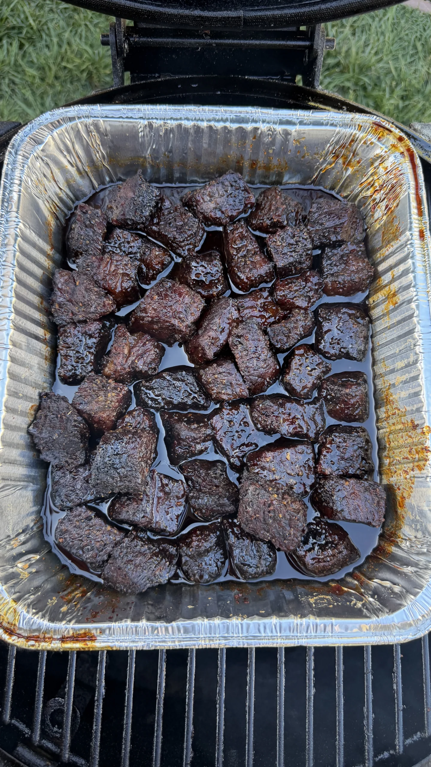 Dark, caramelized smoked molasses coffee beef rib burnt ends in aluminum foil pan on outdoor grill